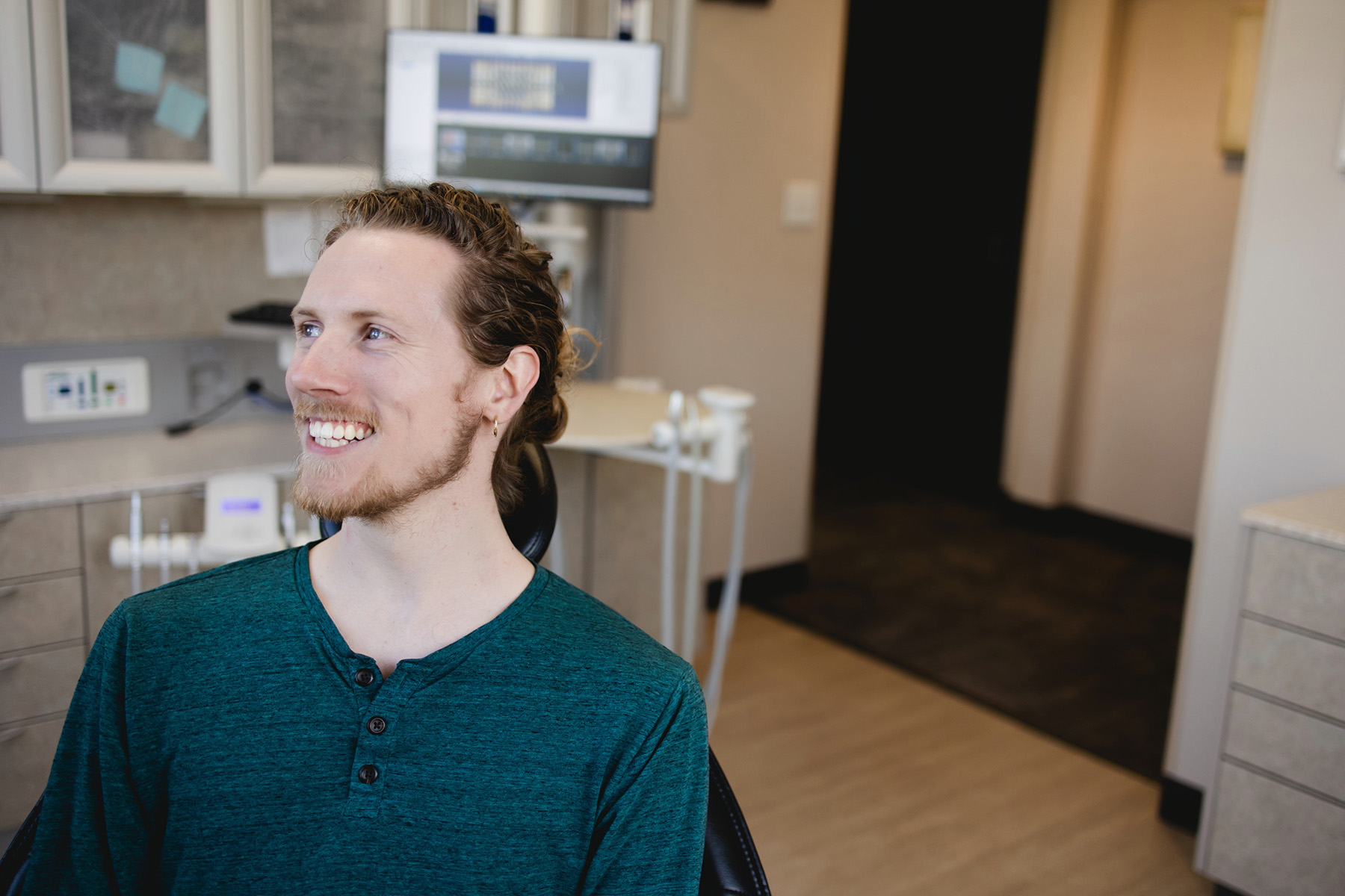 Smiling young man with a beard and curly hair sitting in a dental office chair.