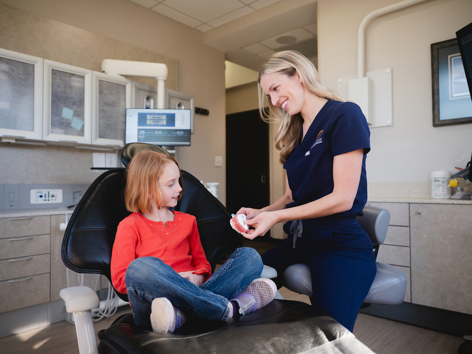 Dentist showing a dental model to a young girl sitting cross-legged on a dental chair in a clinic.