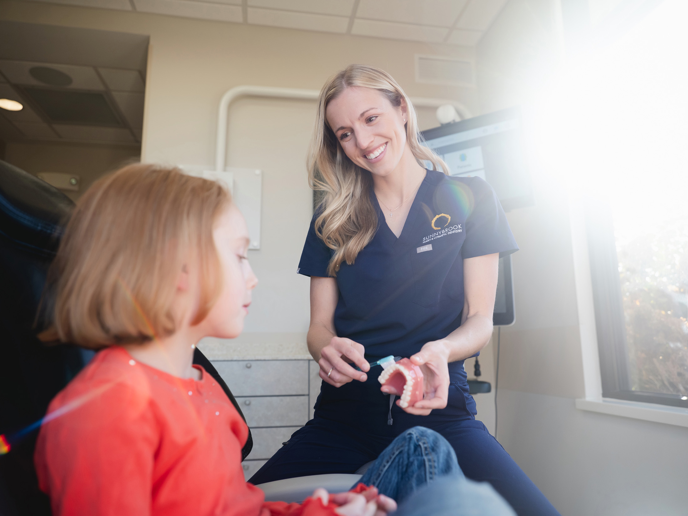 Female dentist in navy scrubs demonstrates brushing on a dental model to a young girl in a dental clinic.