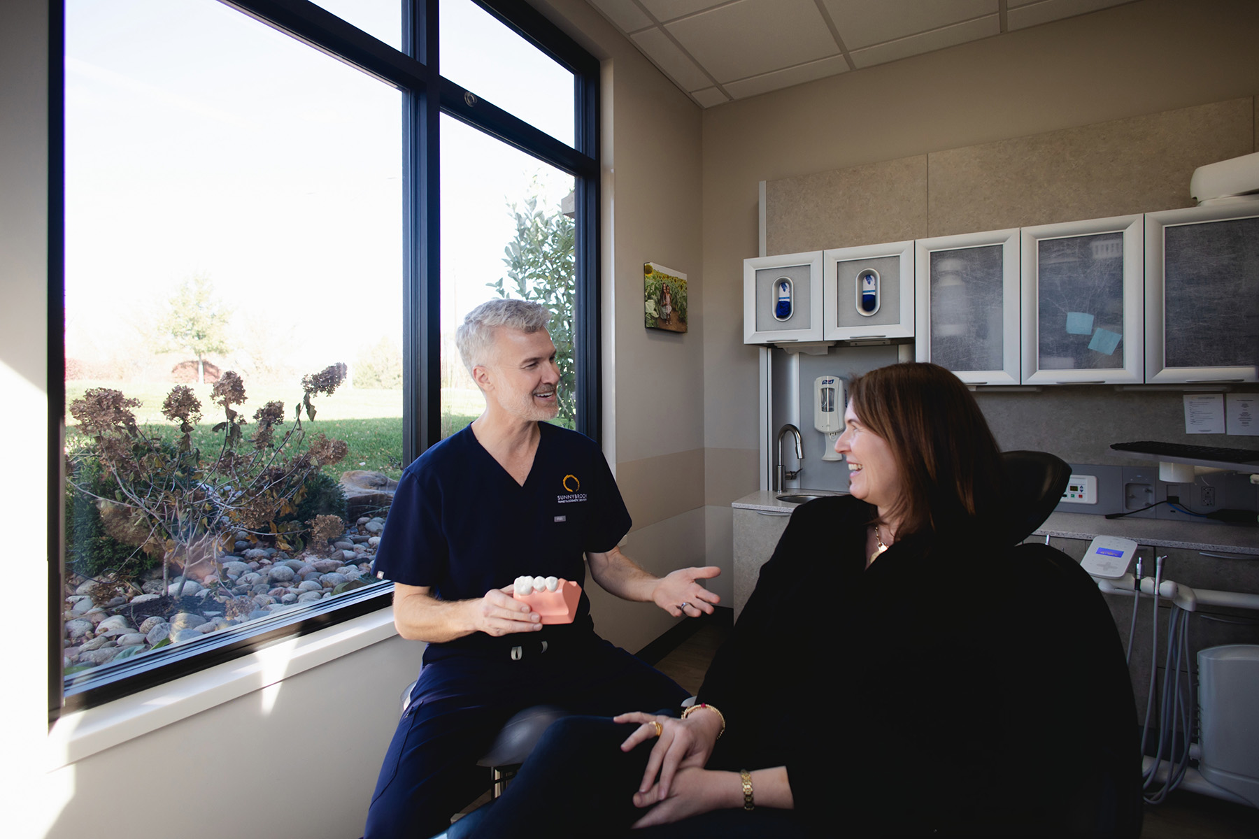 Dentist in navy scrubs holding a dental model and talking to a smiling female patient in a dental office.