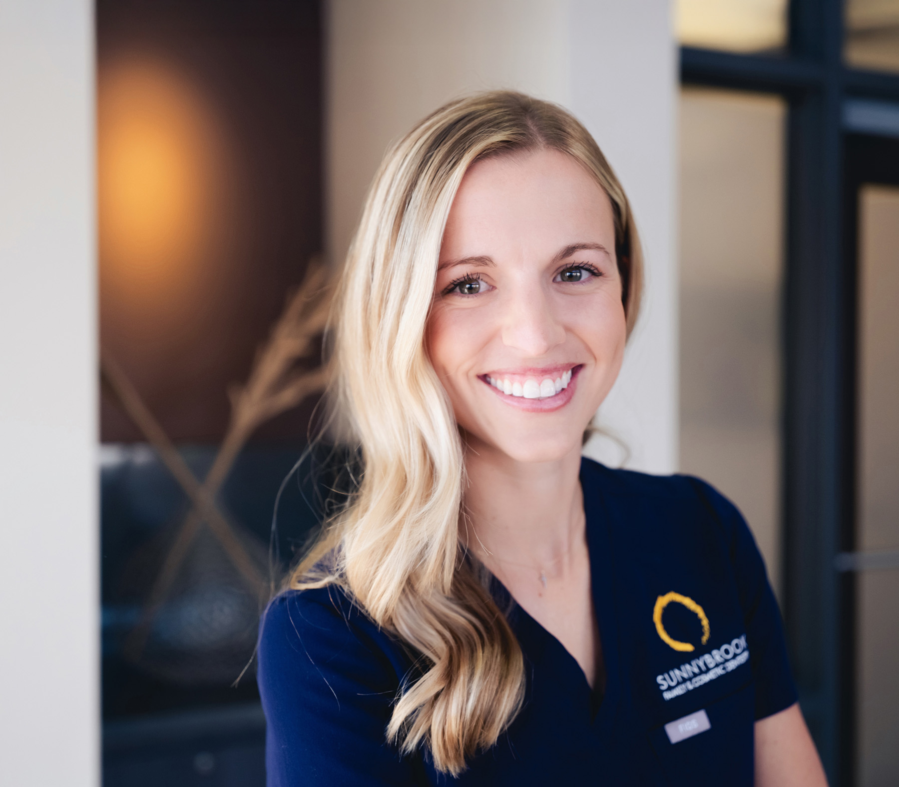Smiling woman with long blonde hair wearing a navy blue shirt with Sunnybrook logo in an indoor setting.