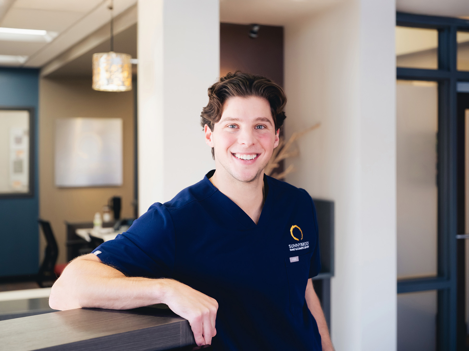 Smiling young man in navy blue Sunnybrook Family Cosmetic Dental scrub standing in a modern office.