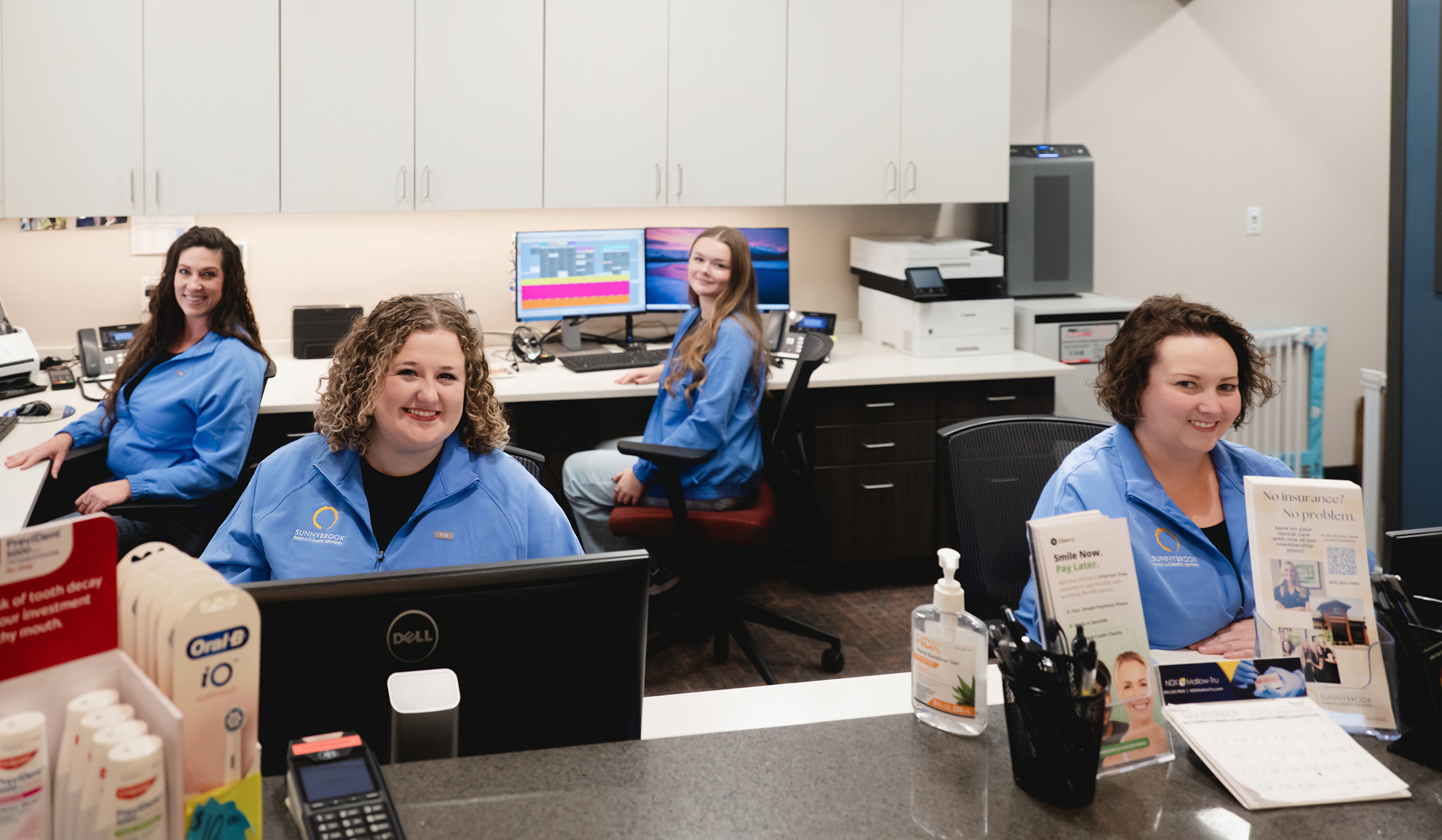 Four women in blue Sunnybrook jackets smiling while sitting at desks in an office reception area.