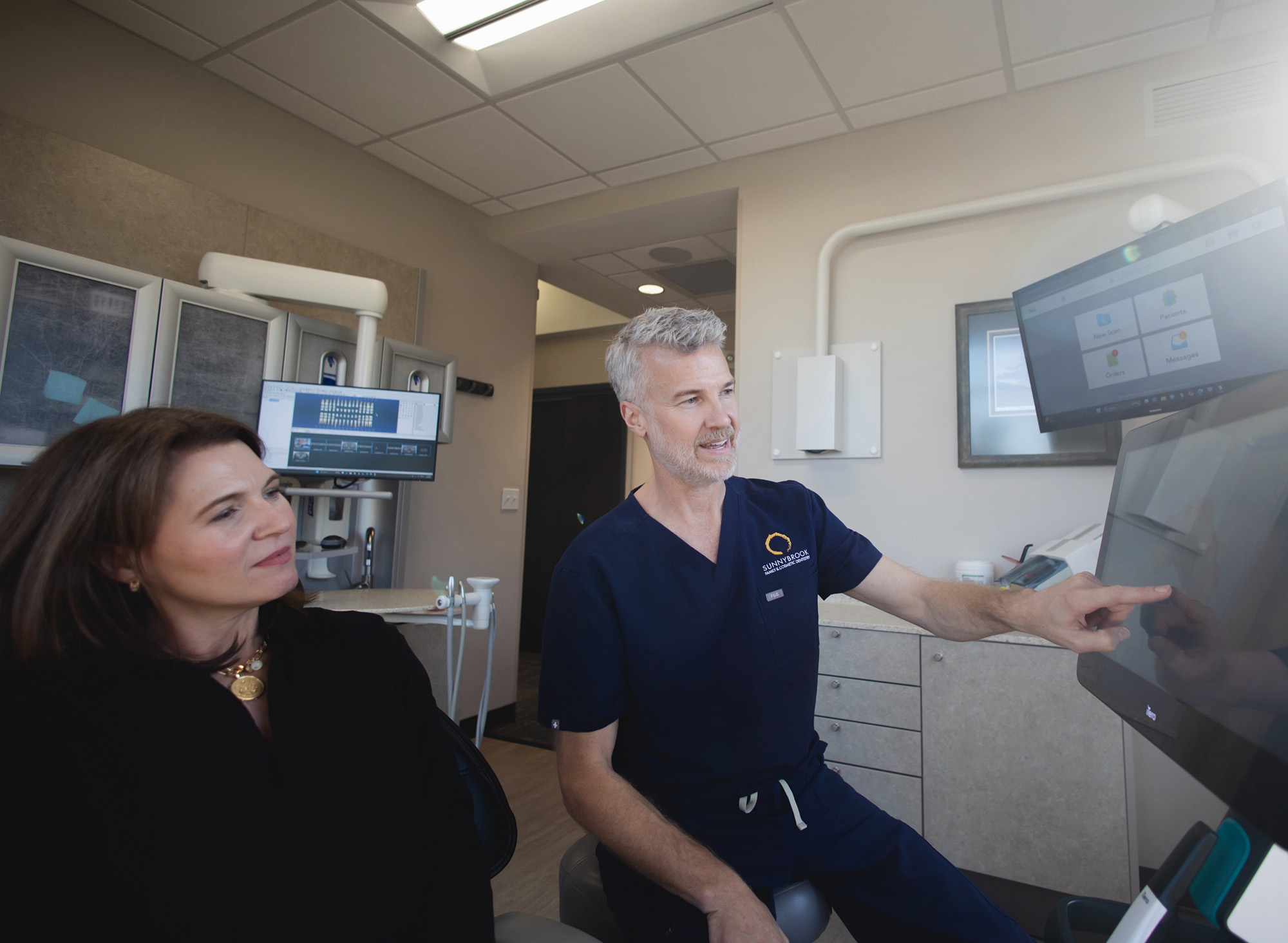 Male dentist in navy scrubs showing a touchscreen display to a female patient in a dental office.