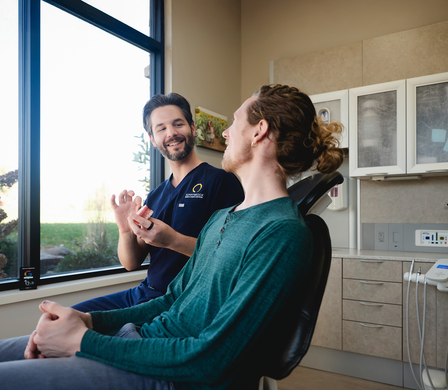 Dentist explaining dentures to a male patient seated in a dental chair by a window in a dental office.