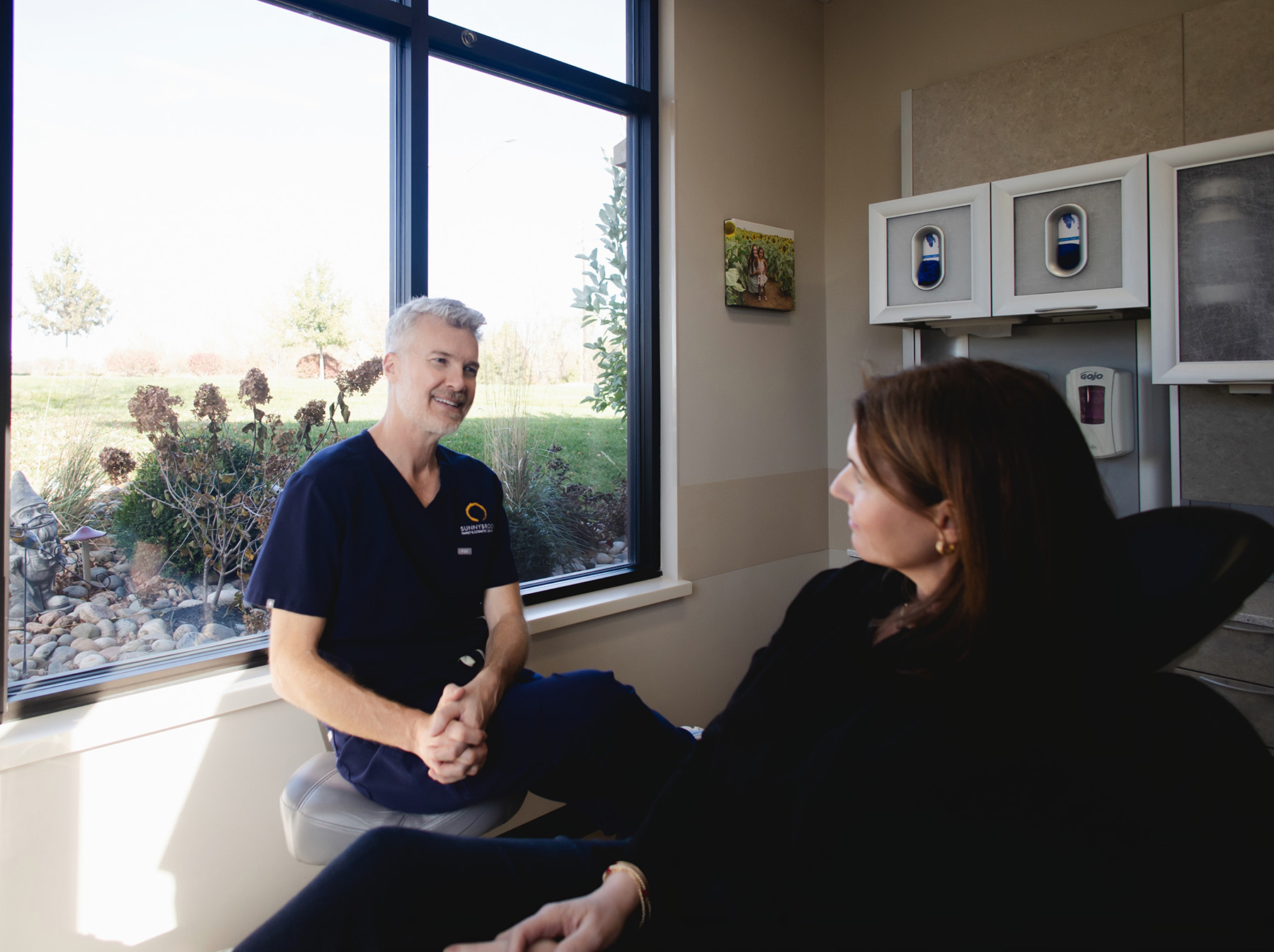 A male dentist in navy scrubs talking with a female patient seated in a dental chair near a large window with a garden view.