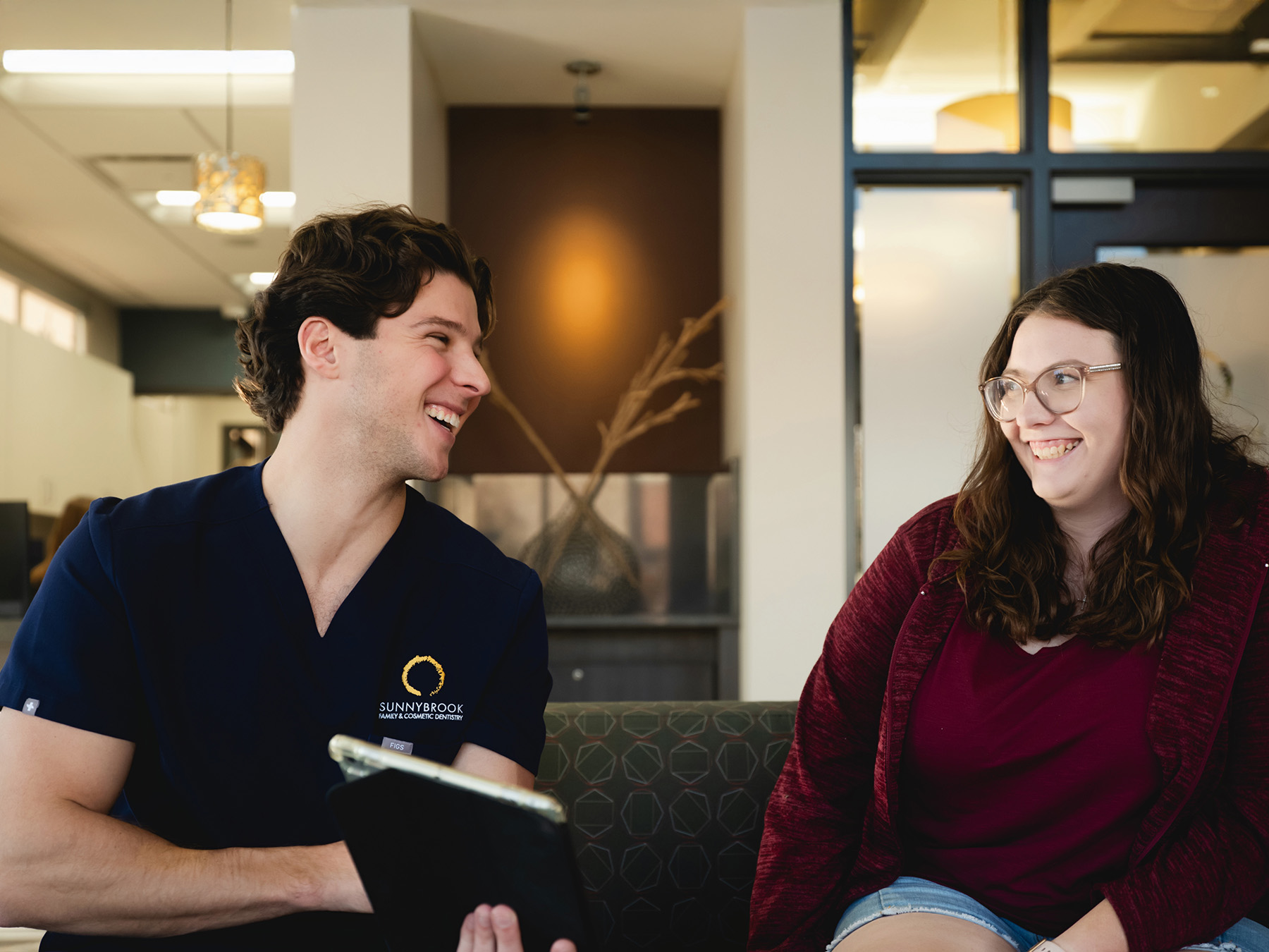 Smiling male dentist in navy scrubs talking to a happy female patient with glasses seated in a waiting room.