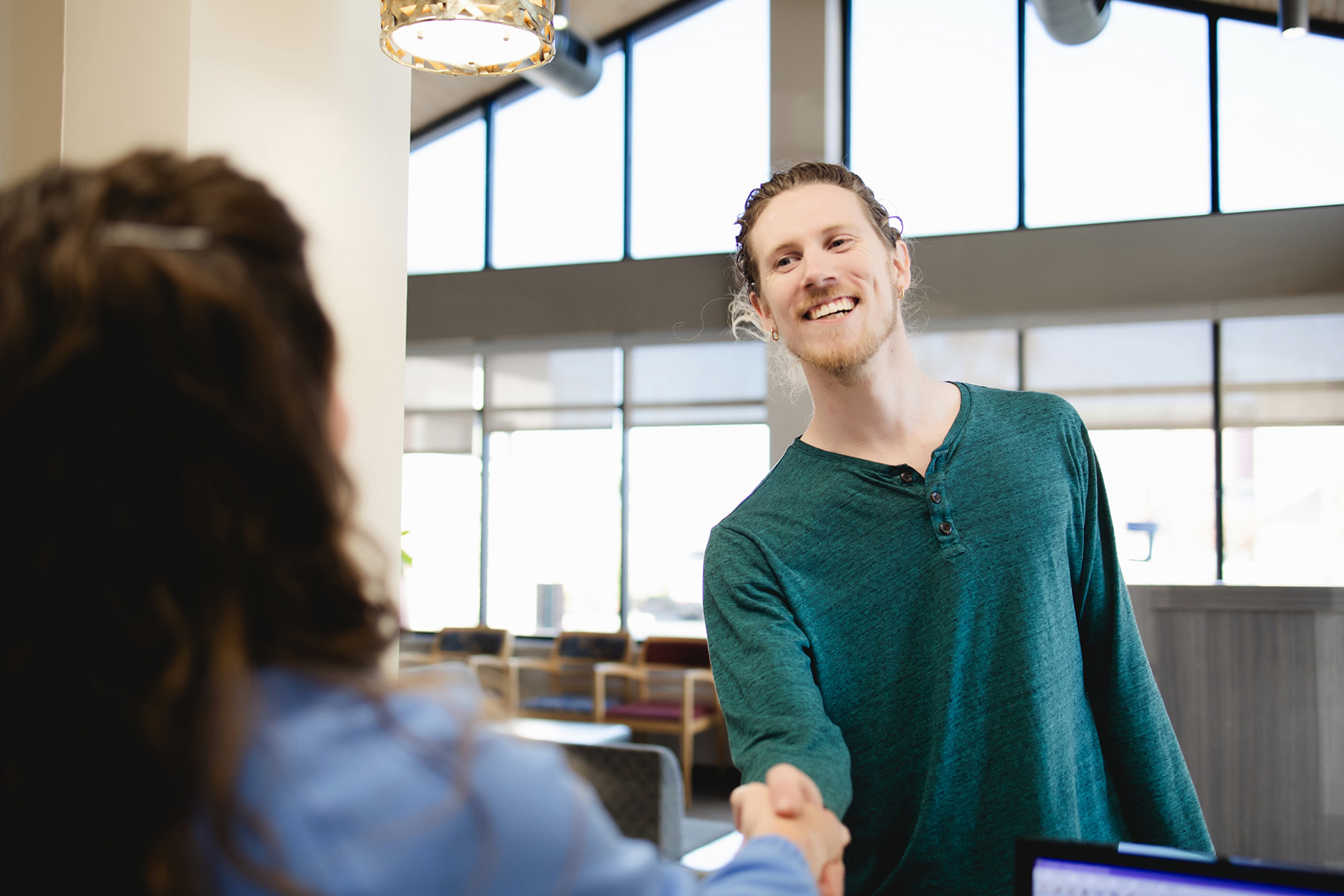 Smiling man in a teal shirt shaking hands with a woman in an office setting with large windows.