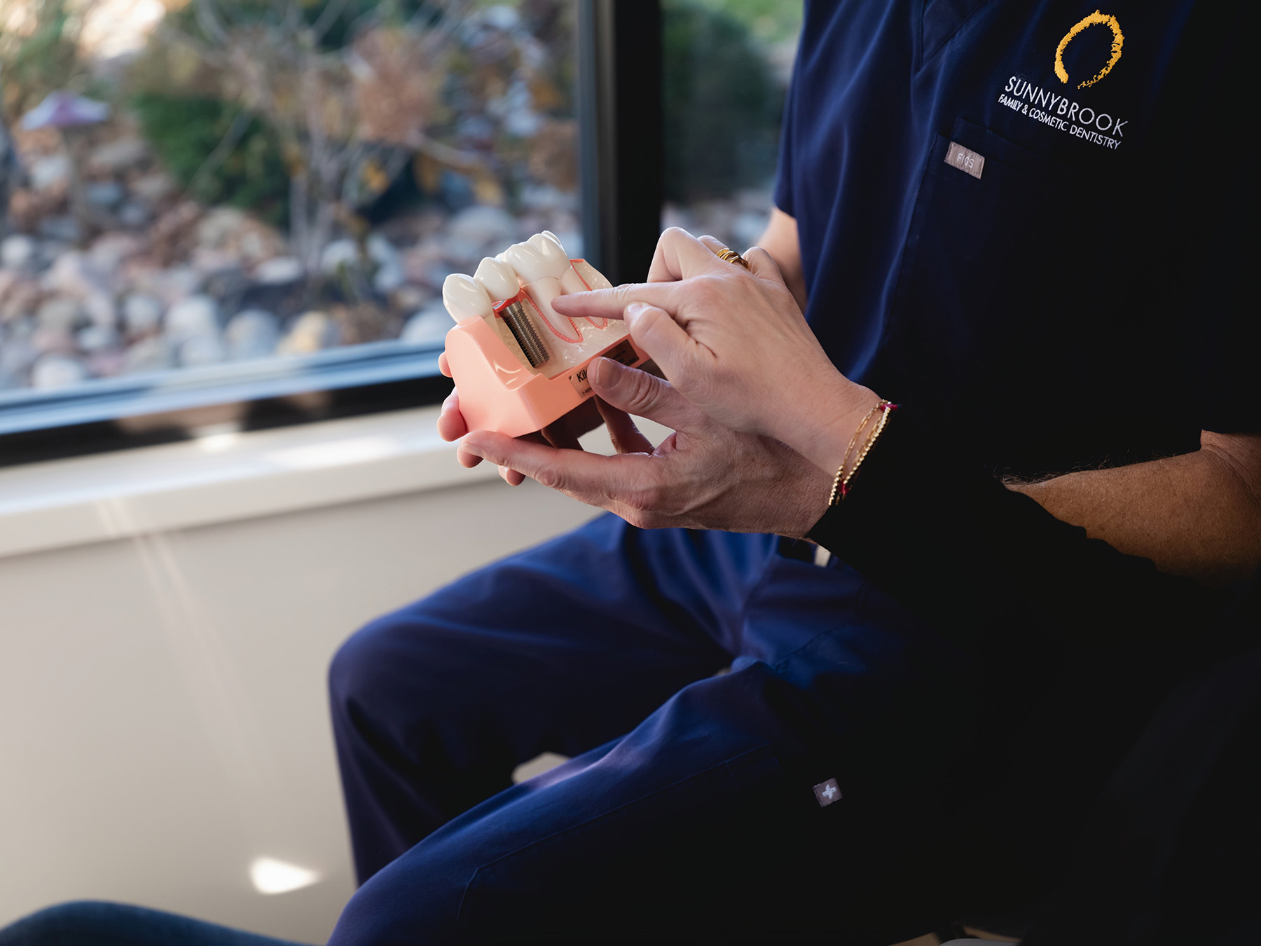 Dentist in navy blue scrubs holding and explaining a dental implant model to a patient.