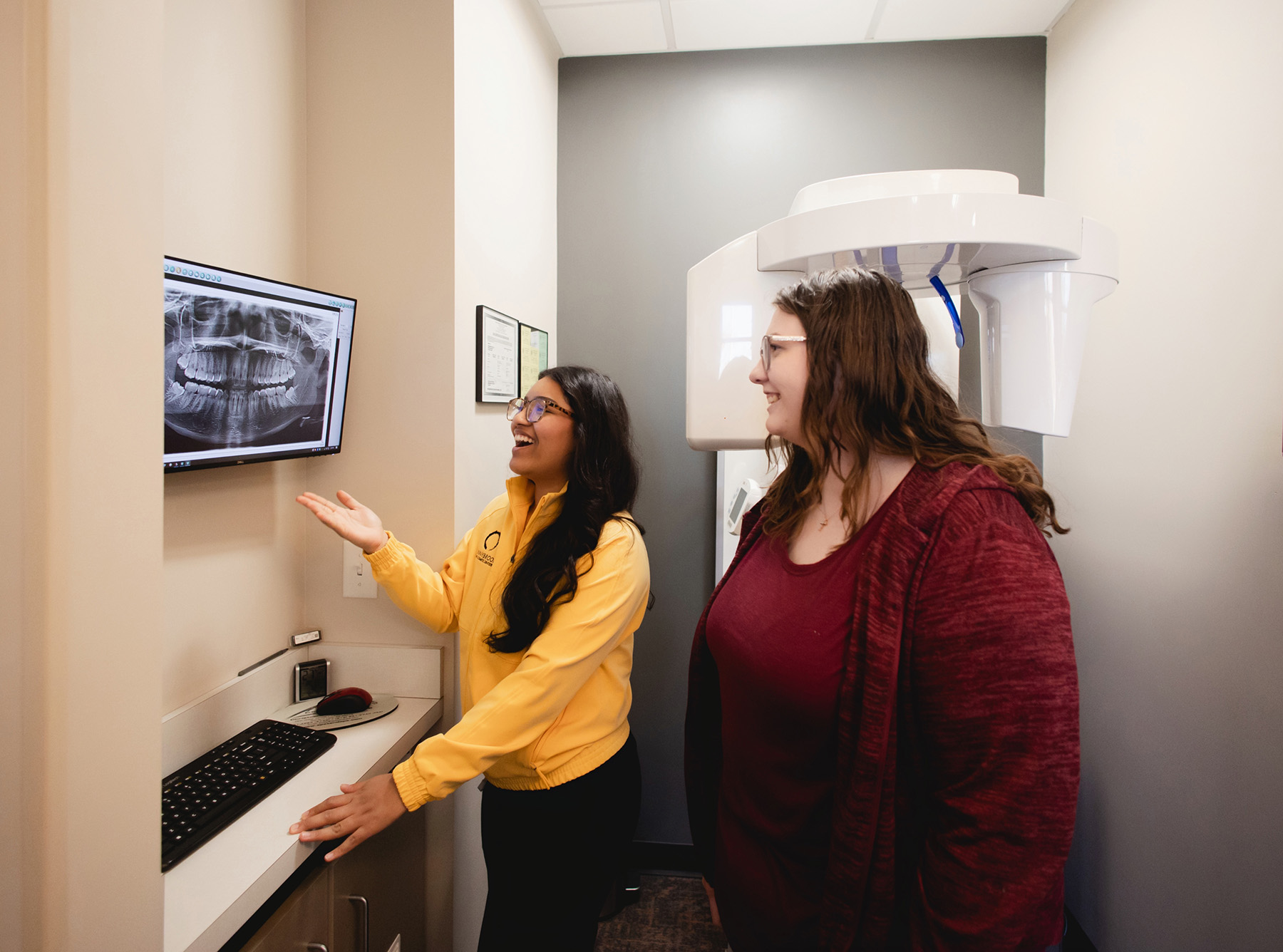 Two women in an X-ray room looking at a dental X-ray displayed on a wall-mounted monitor, with one woman pointing and explaining.