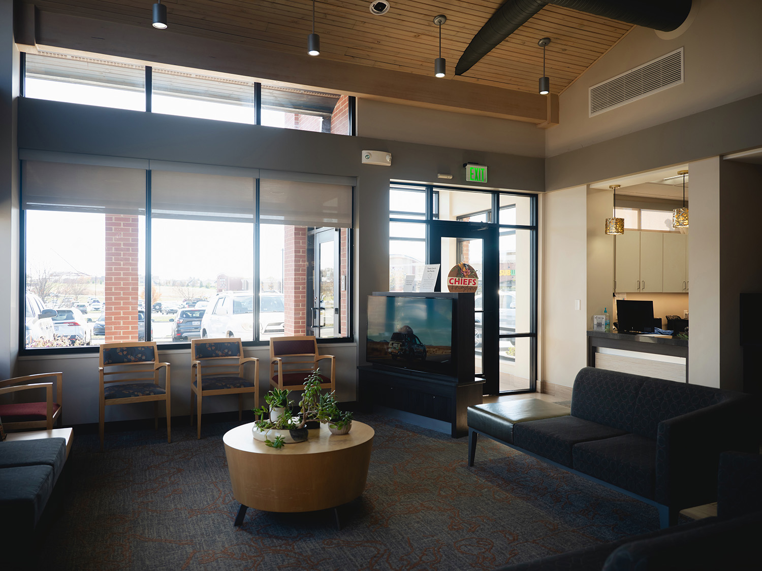 Waiting area with chairs, a round table with potted plants, a television, and a reception desk near large windows and an exit door.