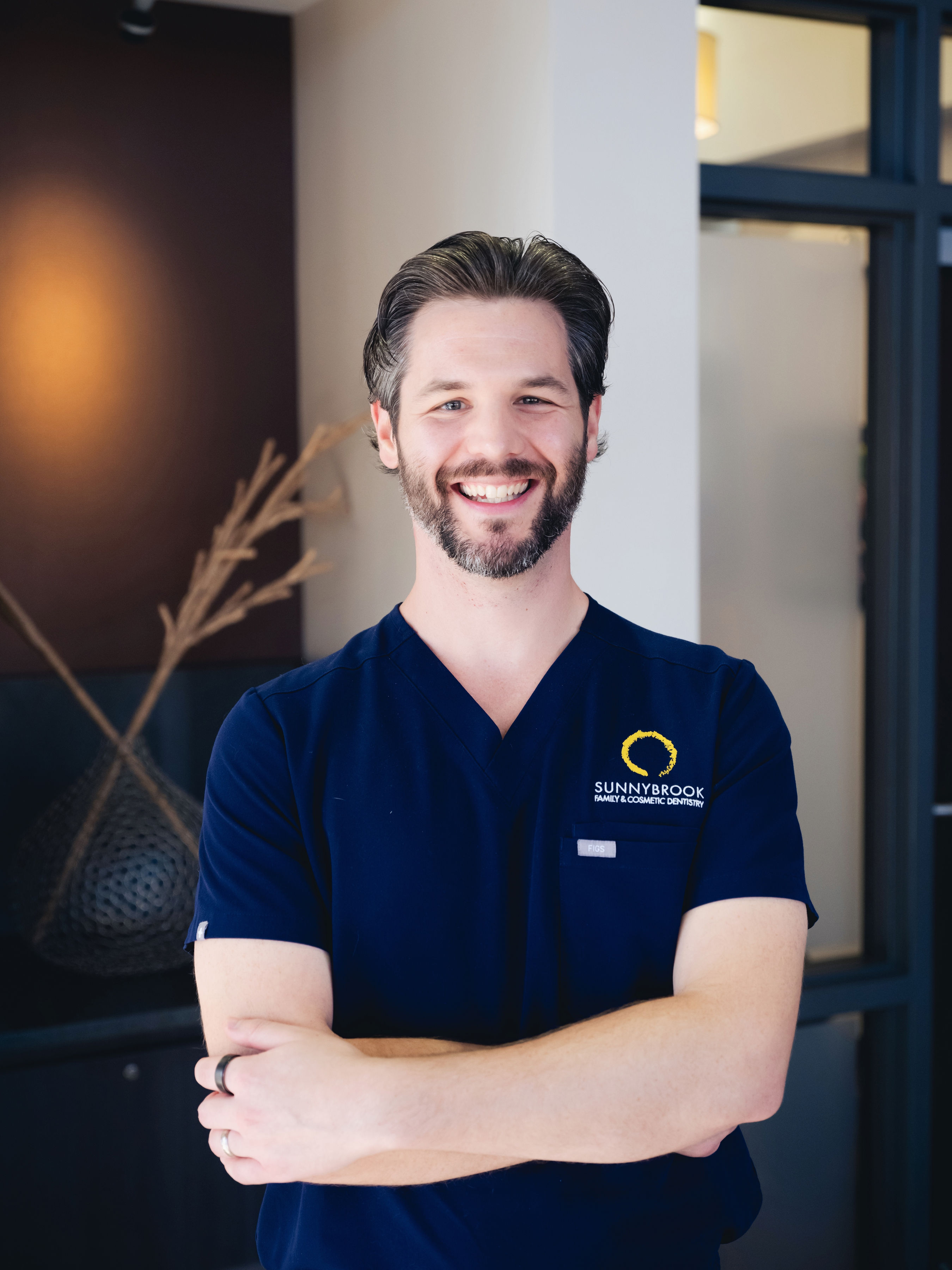 Smiling male dental professional in navy scrubs with Sunnybrook Family & Cosmetic Dentistry logo, standing with arms crossed.