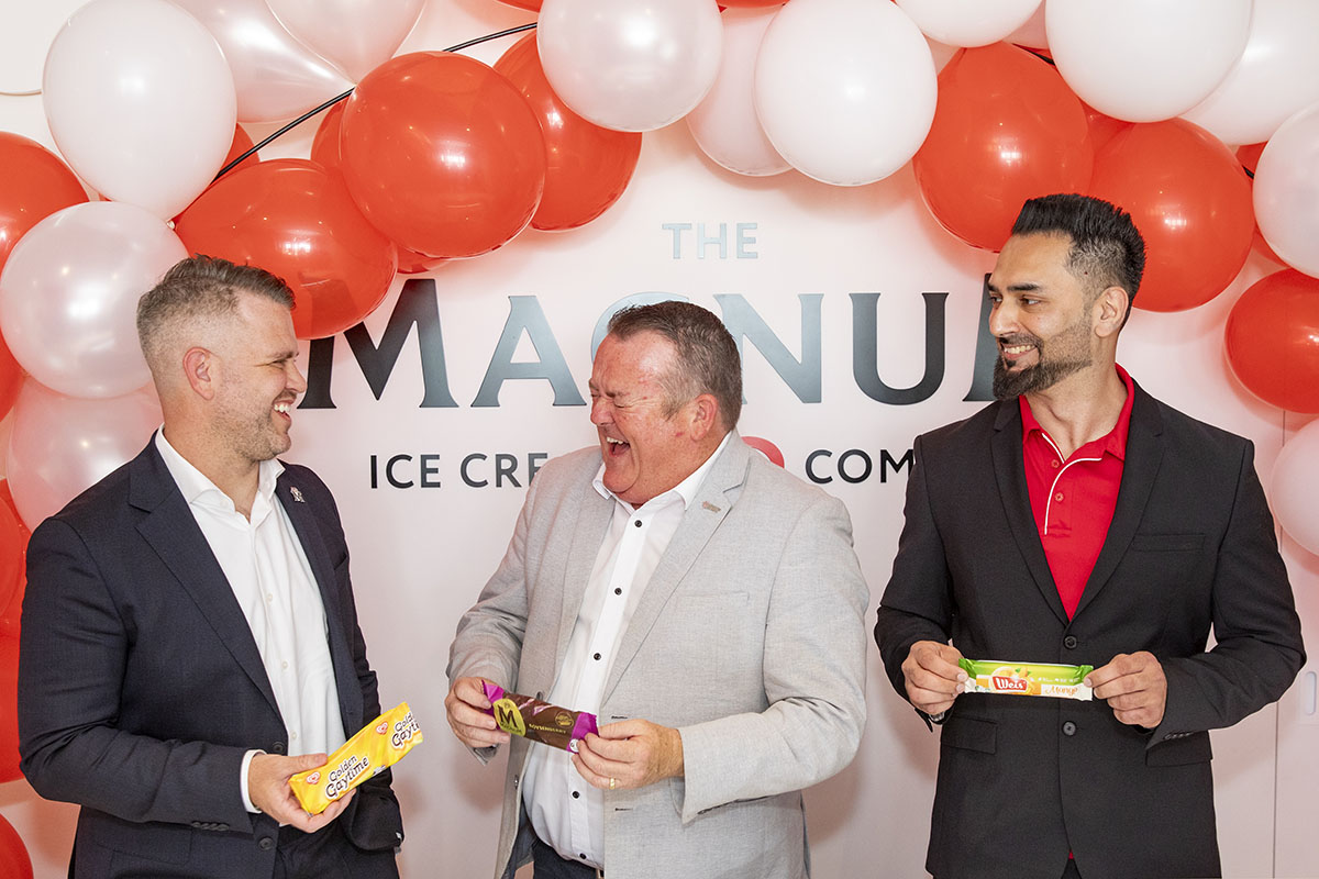 Three men in suits laughing and holding different ice cream bars in front of a red and white balloon arch with 'The Magnum Ice Cream Company' sign.