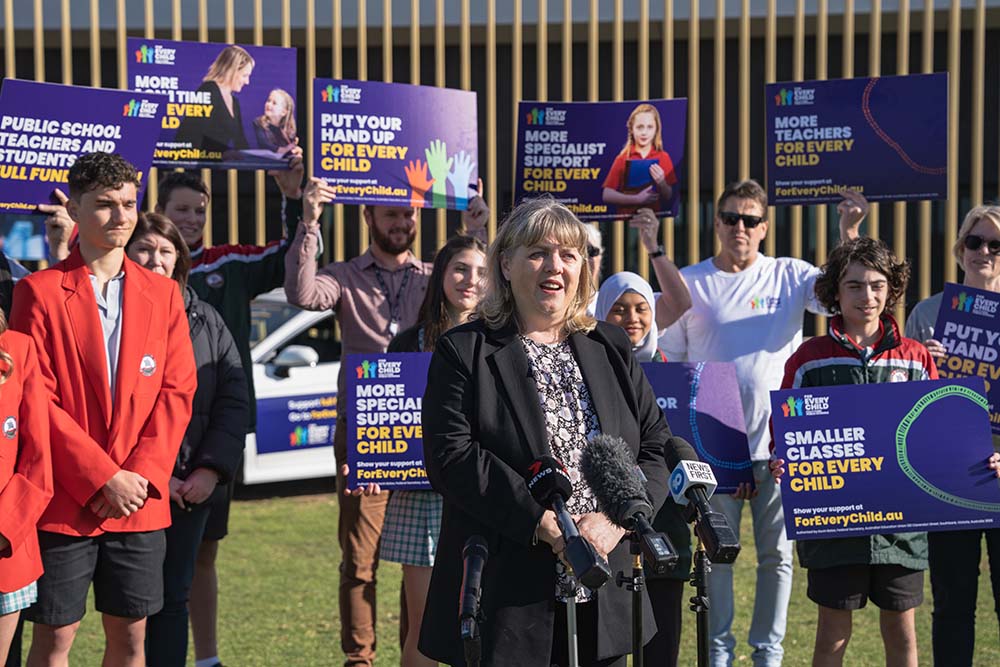 Woman speaking at a press conference outdoors with microphones, surrounded by a diverse group of people holding signs advocating for education support and smaller classes for every child.