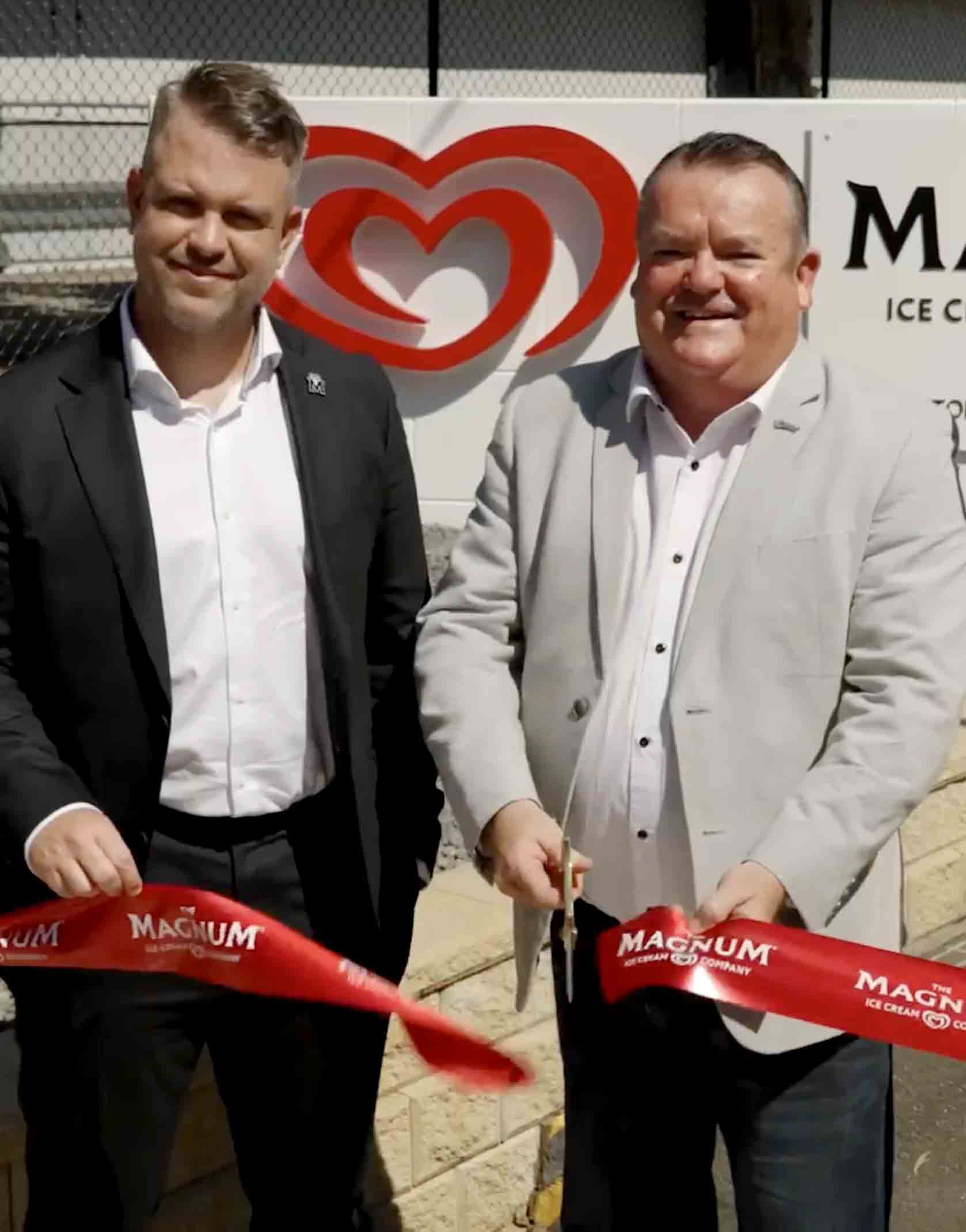 Two men in suits smiling while cutting a red ribbon with the Magnum Ice Cream Company logo at an outdoor event.