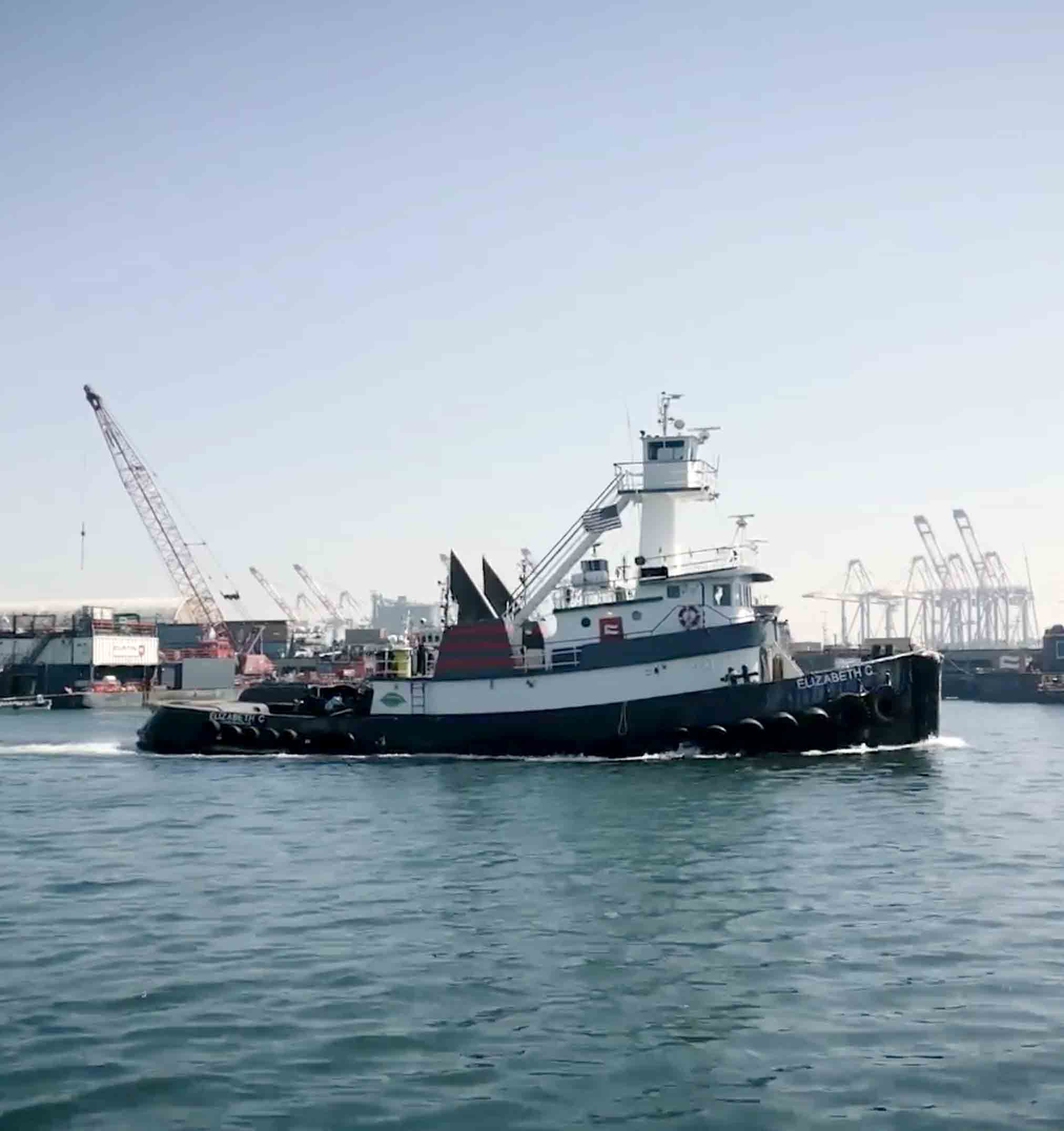 Tugboat named Elizabeth C moving through a harbor with cranes and shipping containers in the background.