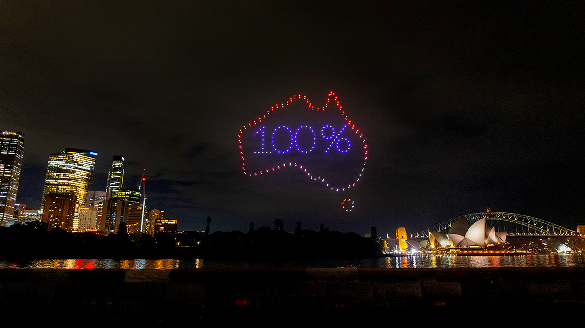 Night skyline of Sydney with illuminated buildings, Sydney Opera House, Harbour Bridge, and a drone light display showing '100%' inside an outline of Australia.