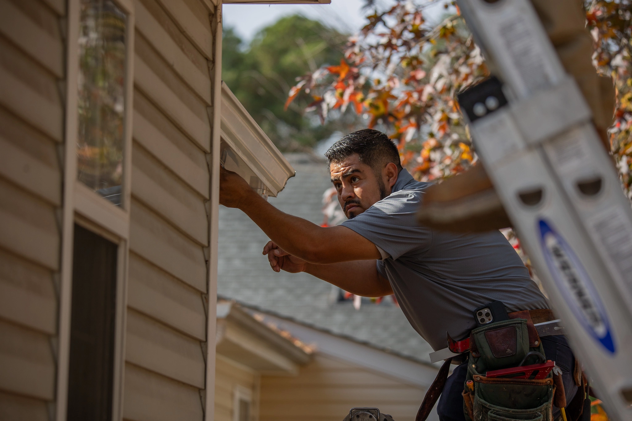 Worker installing or repairing white gutter on beige house siding, standing beside a ladder.