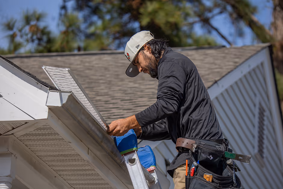 Man on ladder installing a gutter guard on a house gutter under clear sky.