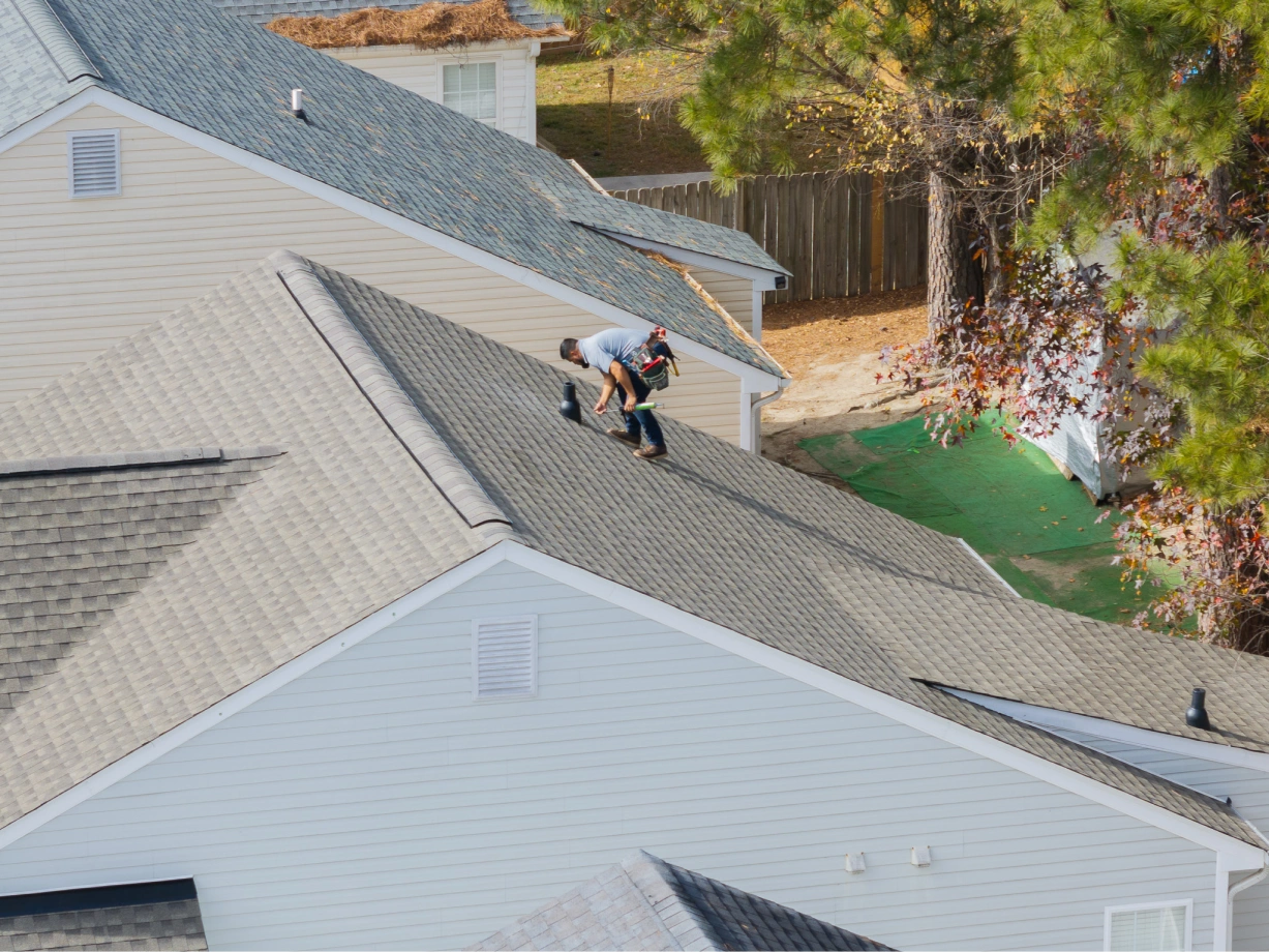Worker wearing a tool belt inspecting or repairing the roof of a house with gray shingles.