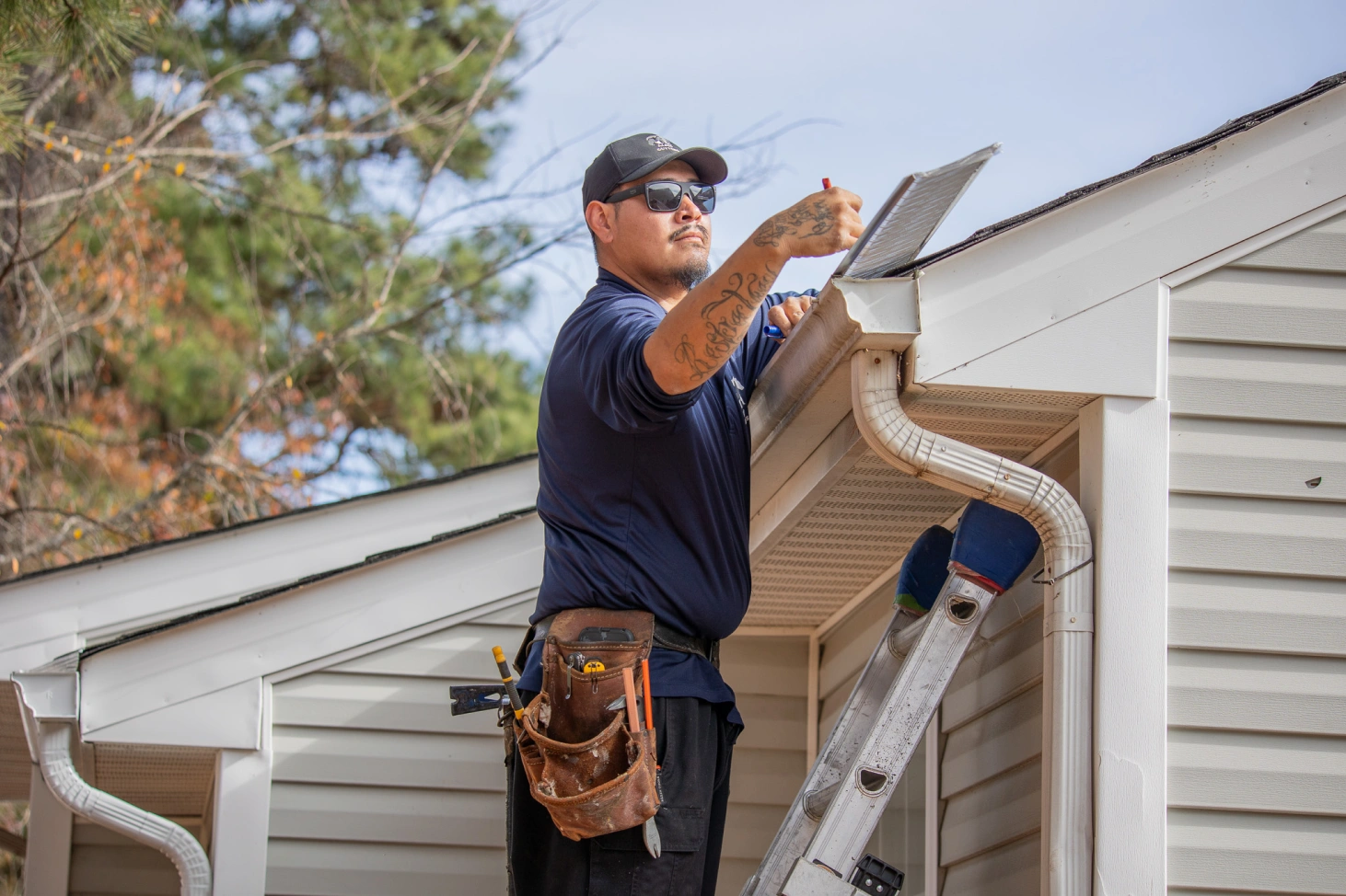 Man wearing sunglasses and a cap inspecting and cleaning a home's gutter while standing on a ladder.