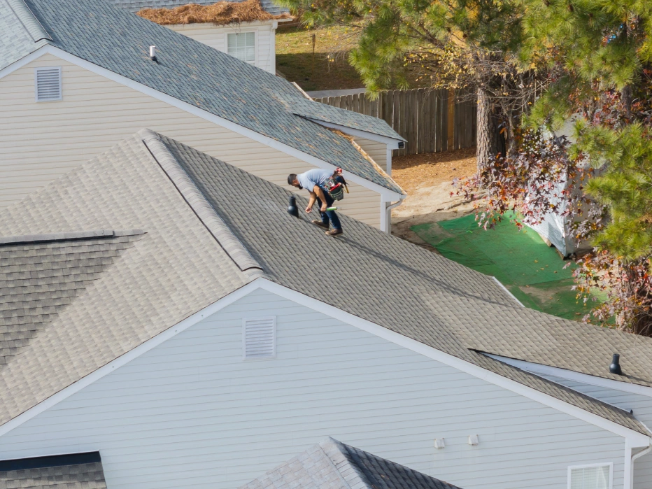 A worker wearing a tool belt is kneeling on a gray shingled roof of a residential house during daytime.