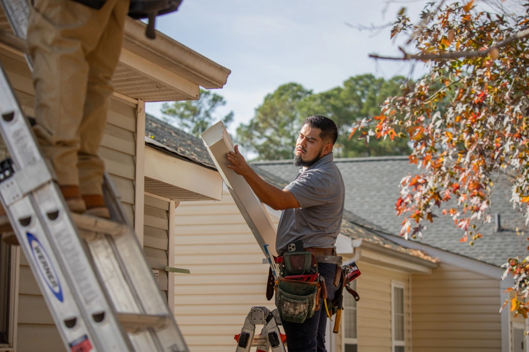 Construction worker measuring a white board while standing on a ladder near a beige house on a sunny day.