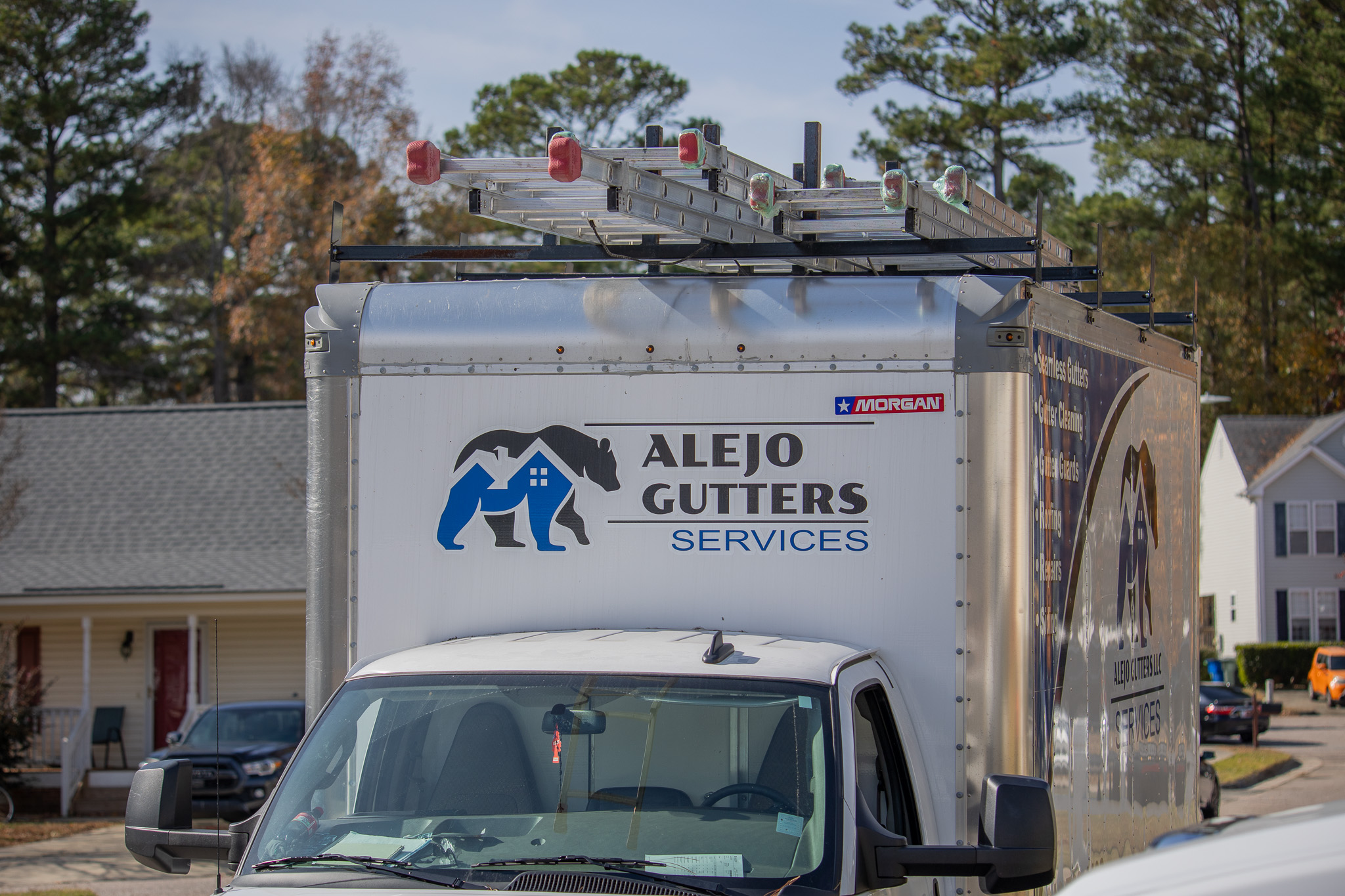 White service truck with Alejo Gutters Services logo and ladders mounted on top, parked in a residential neighborhood.