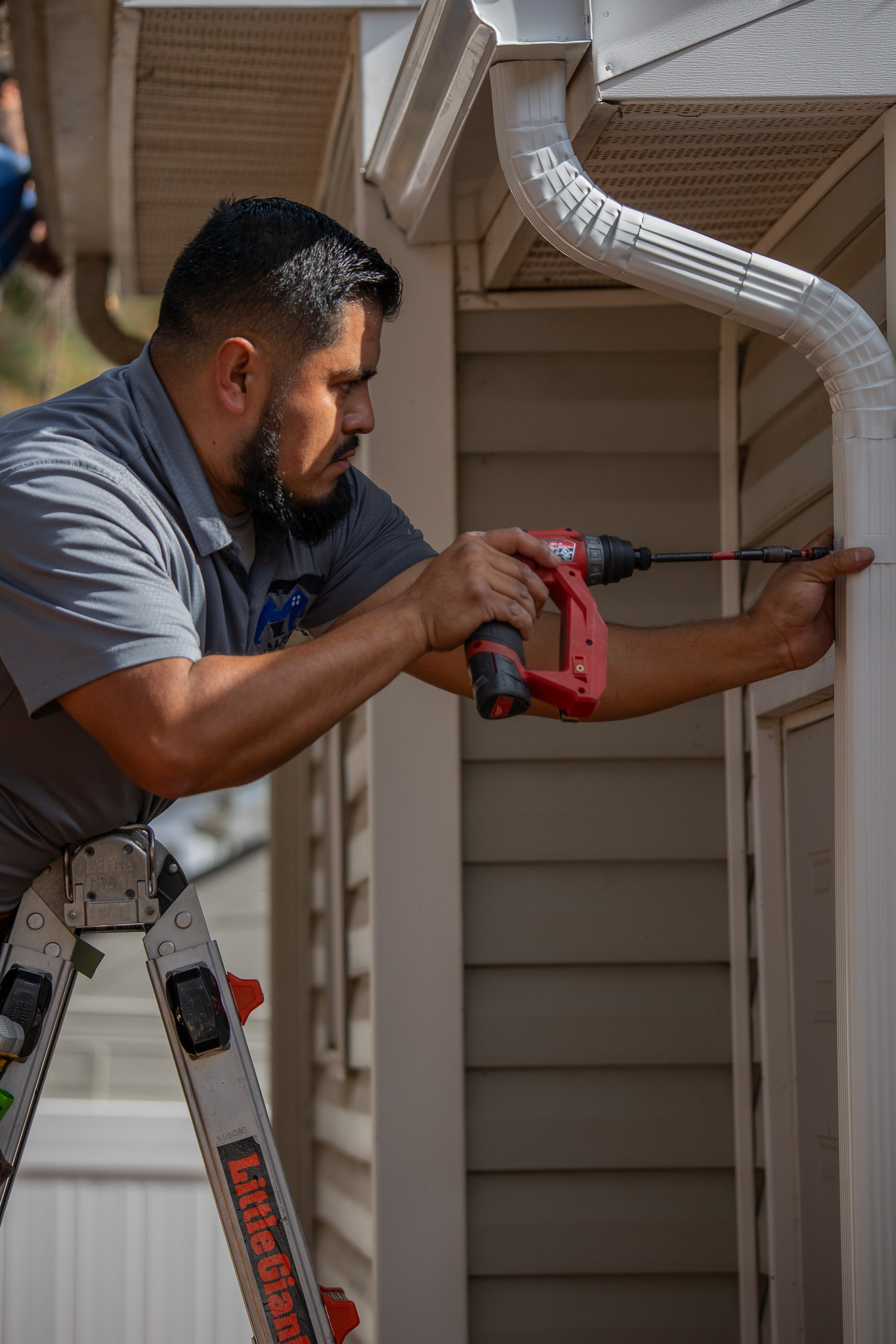 Man on ladder using a red power drill to install or fix a white downspout on a house exterior.