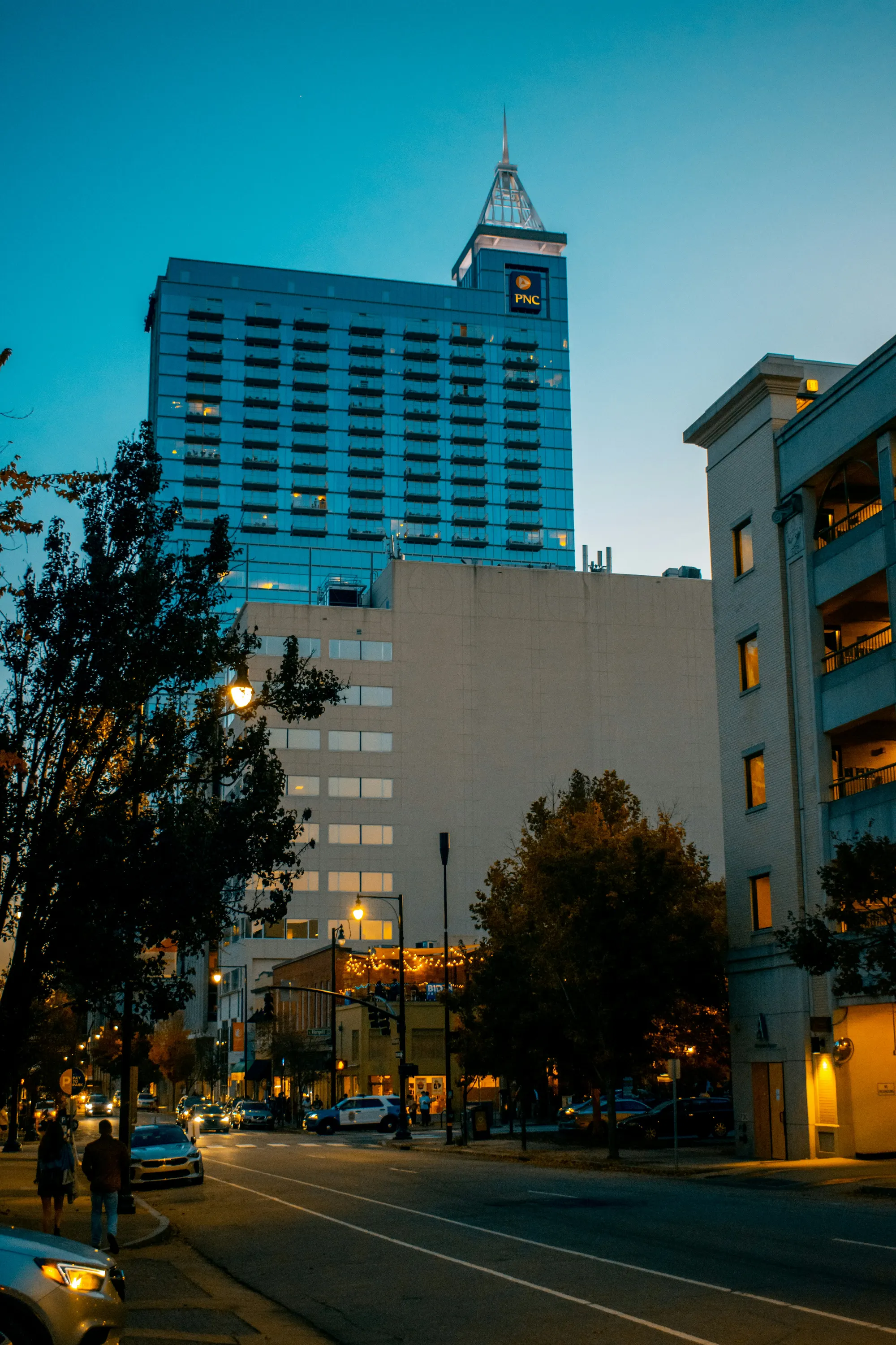 Evening street view in a city with a tall glass building displaying a PNC logo and lit streetlights.