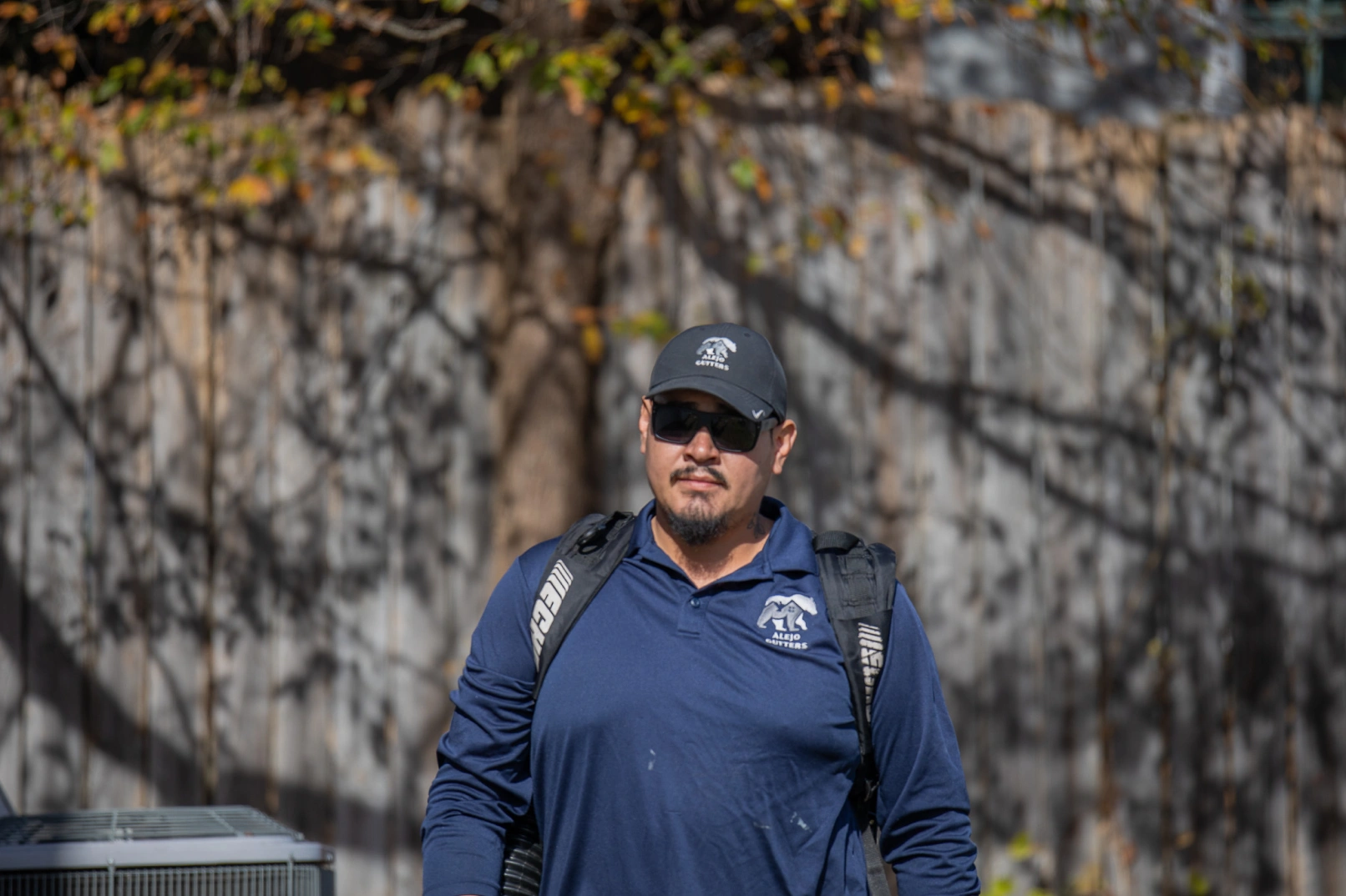 Man wearing sunglasses, navy blue shirt and cap with Alejo Gutters logo walking outside near a fence with tree shadows.