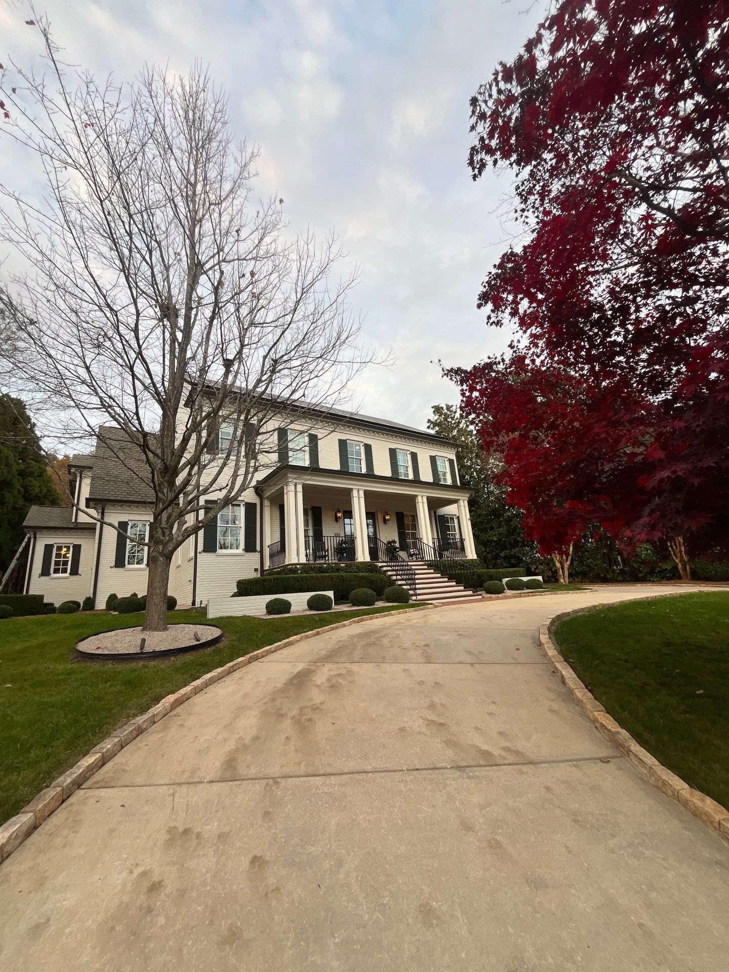 Two-story white house with black shutters, a columned porch, curved driveway, leafless tree on the left, and a red-leaved tree on the right.