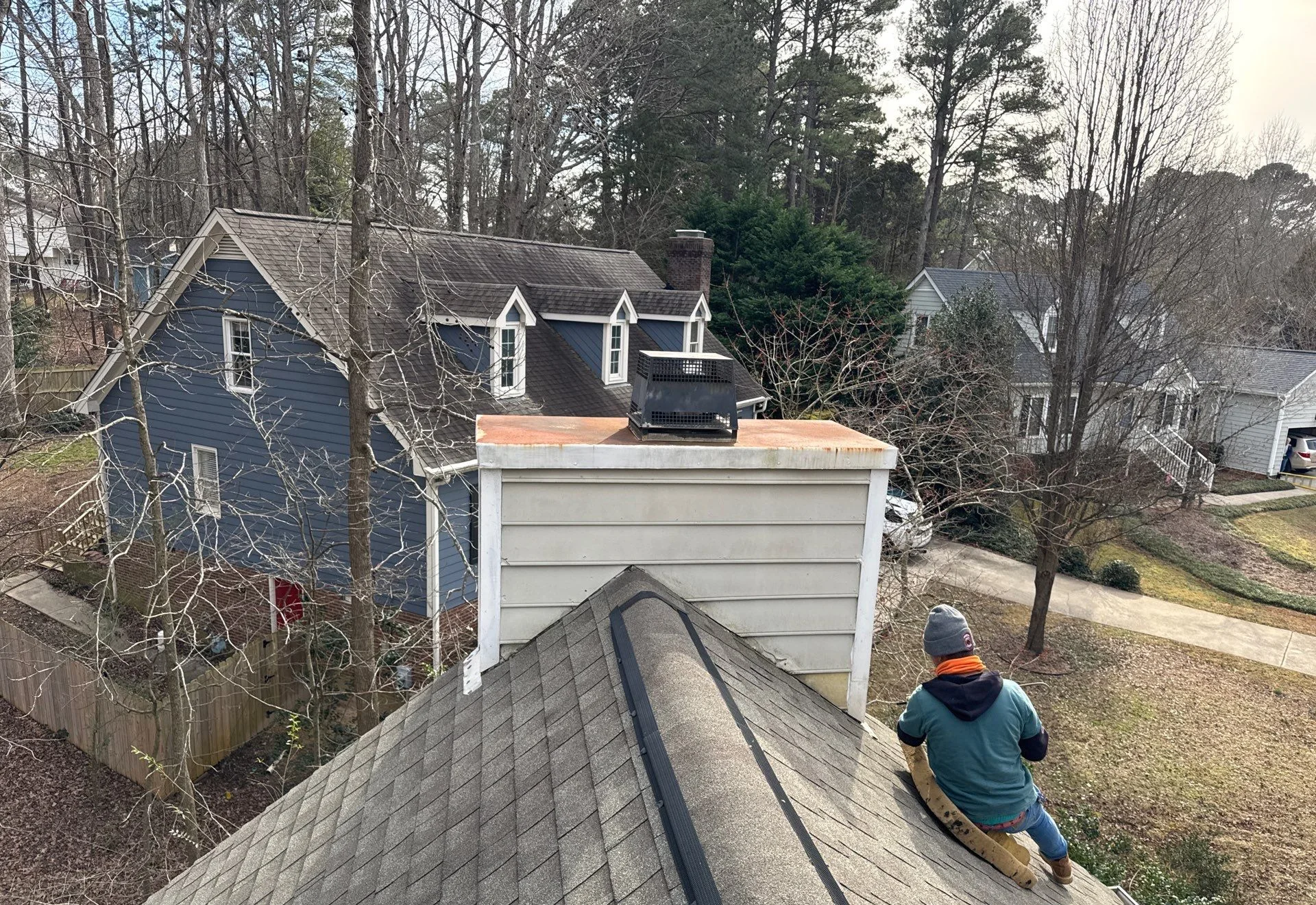 Worker sitting on a roof near a chimney in a residential neighborhood with houses and bare trees.