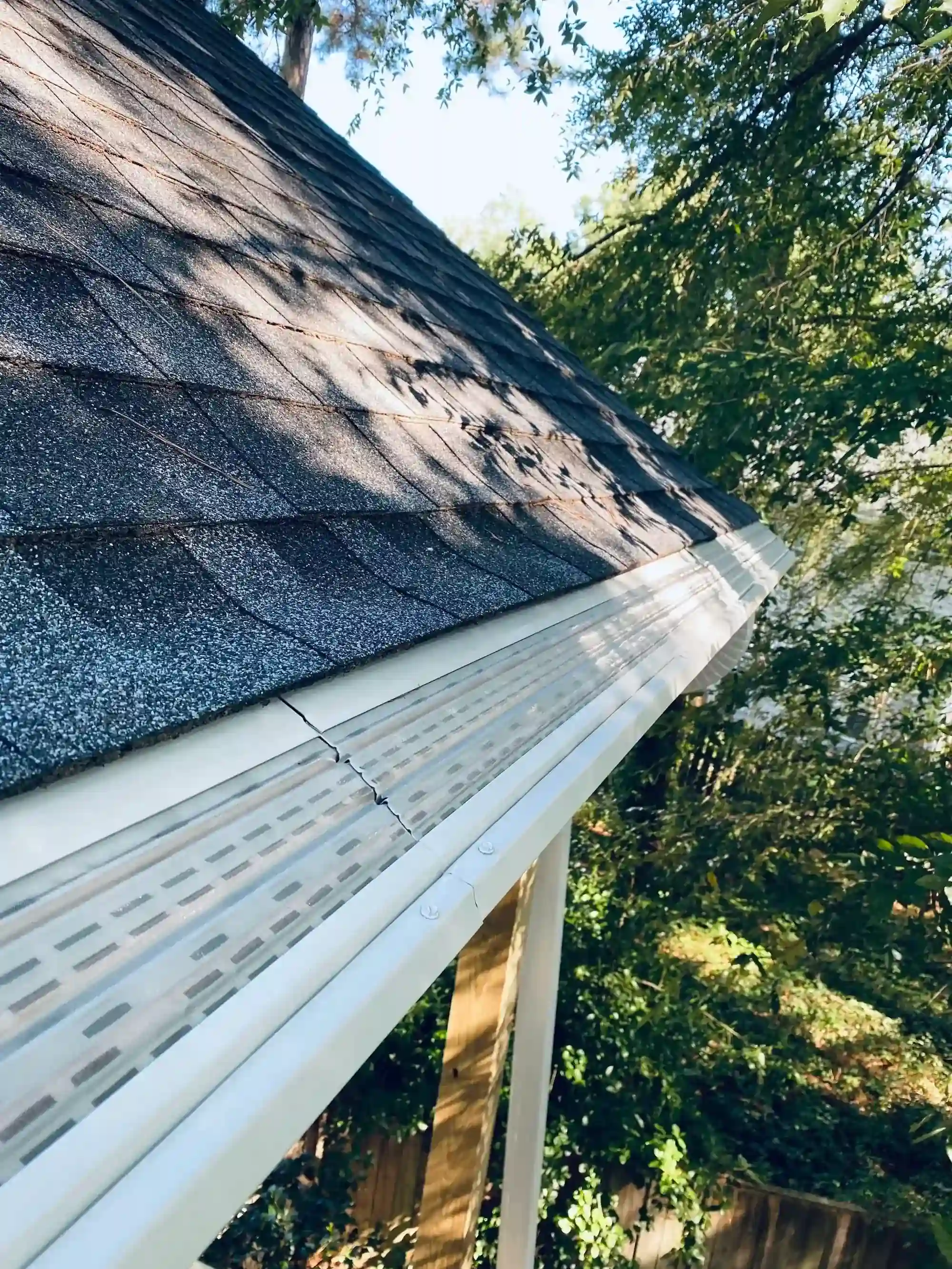 Close-up of a house roof with gray asphalt shingles and a clean white gutter with a leaf guard, surrounded by green trees.