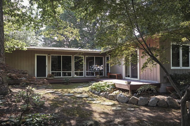 Single-story house with large windows and a wooden deck surrounded by trees and a stone-bordered garden pathway.