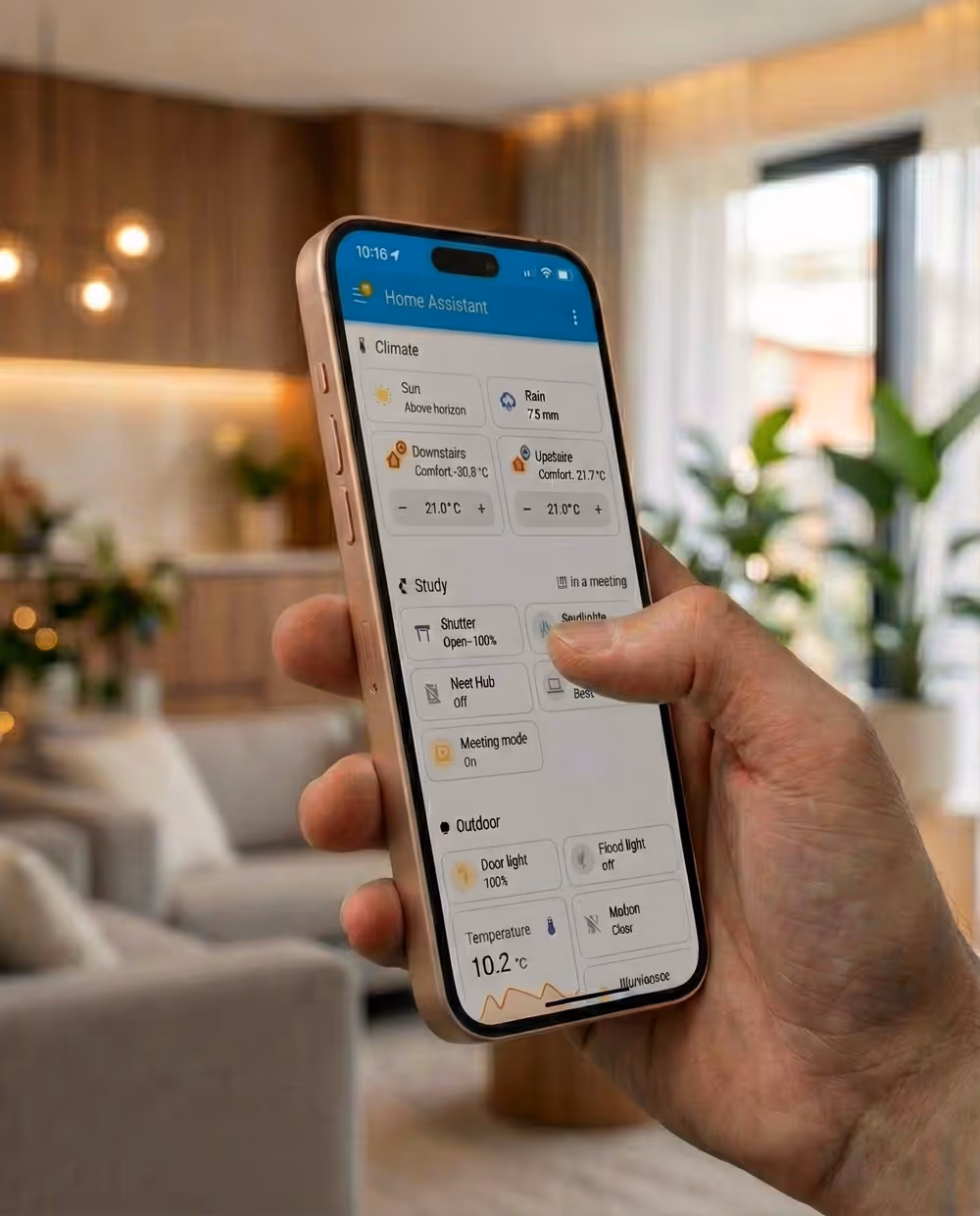 Hand holding smartphone displaying a home assistant app with climate, study, and outdoor controls in a cozy living room.