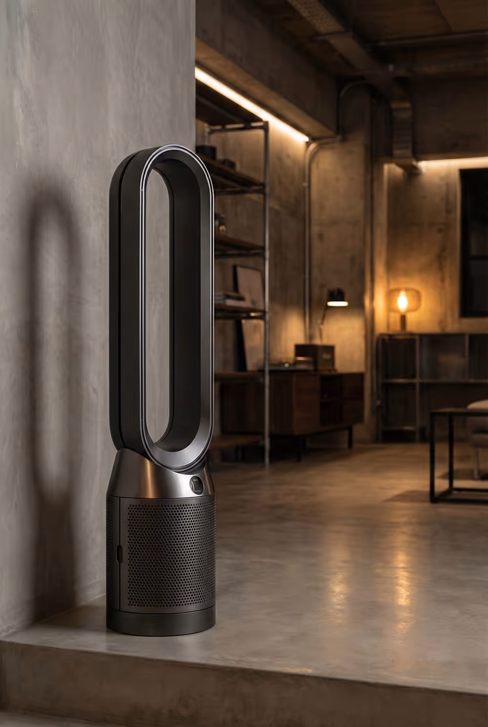 Modern bladeless tower fan placed on a concrete floor in an industrial-style room with dim warm lighting.