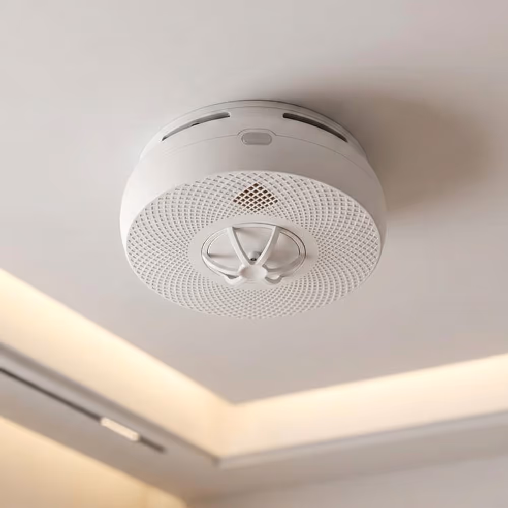 White smoke detector mounted on a ceiling near recessed lighting.