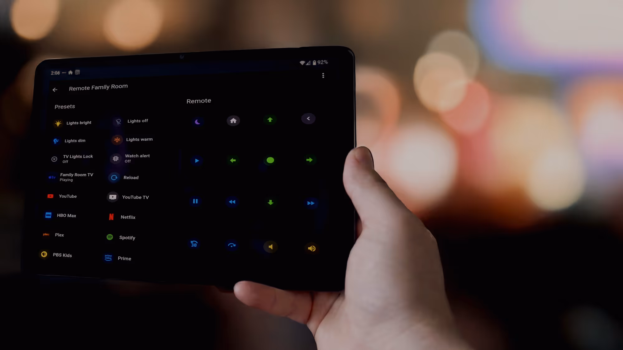 Hand holding a tablet displaying a smart home app with presets and remote control for family room lights and streaming services.