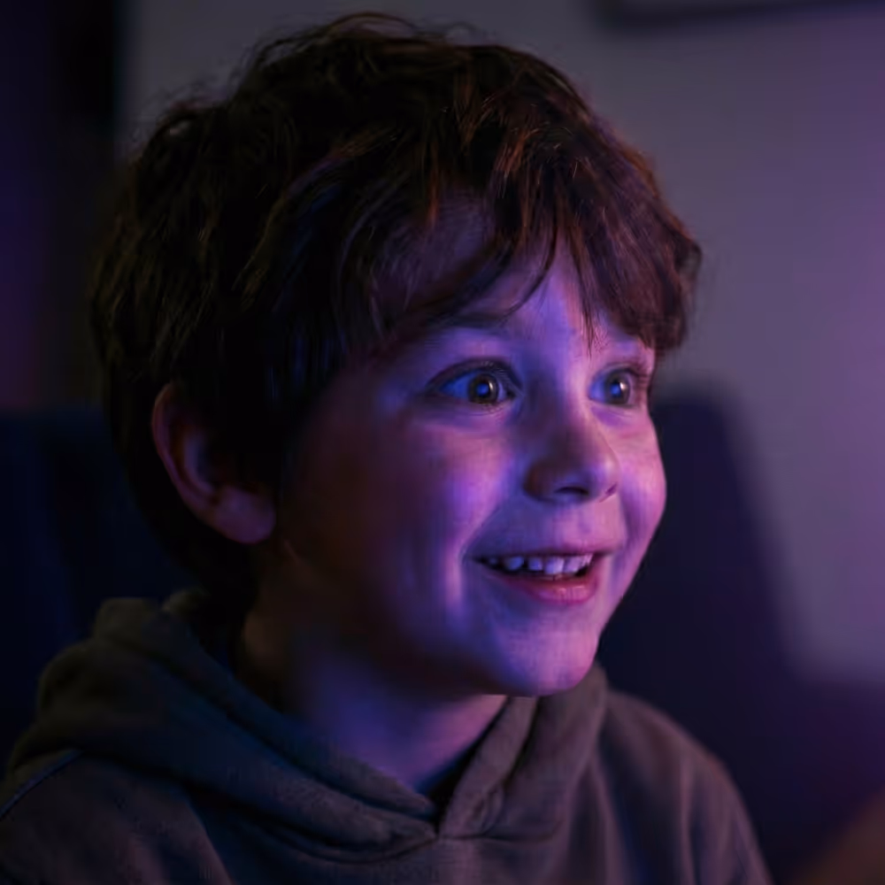 Excited young boy with curly hair illuminated by purple light, smiling and looking ahead.