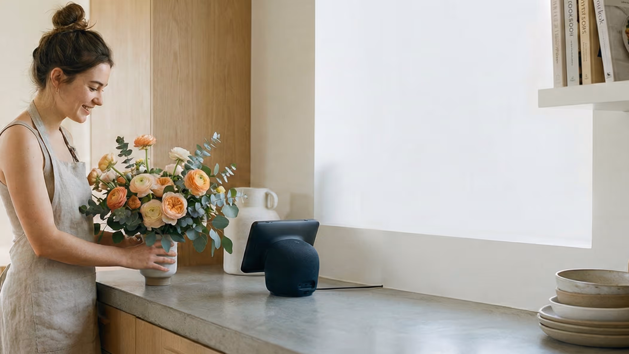 Woman arranging a bouquet of orange and white flowers in a vase on a kitchen countertop near a smart speaker and tablet.