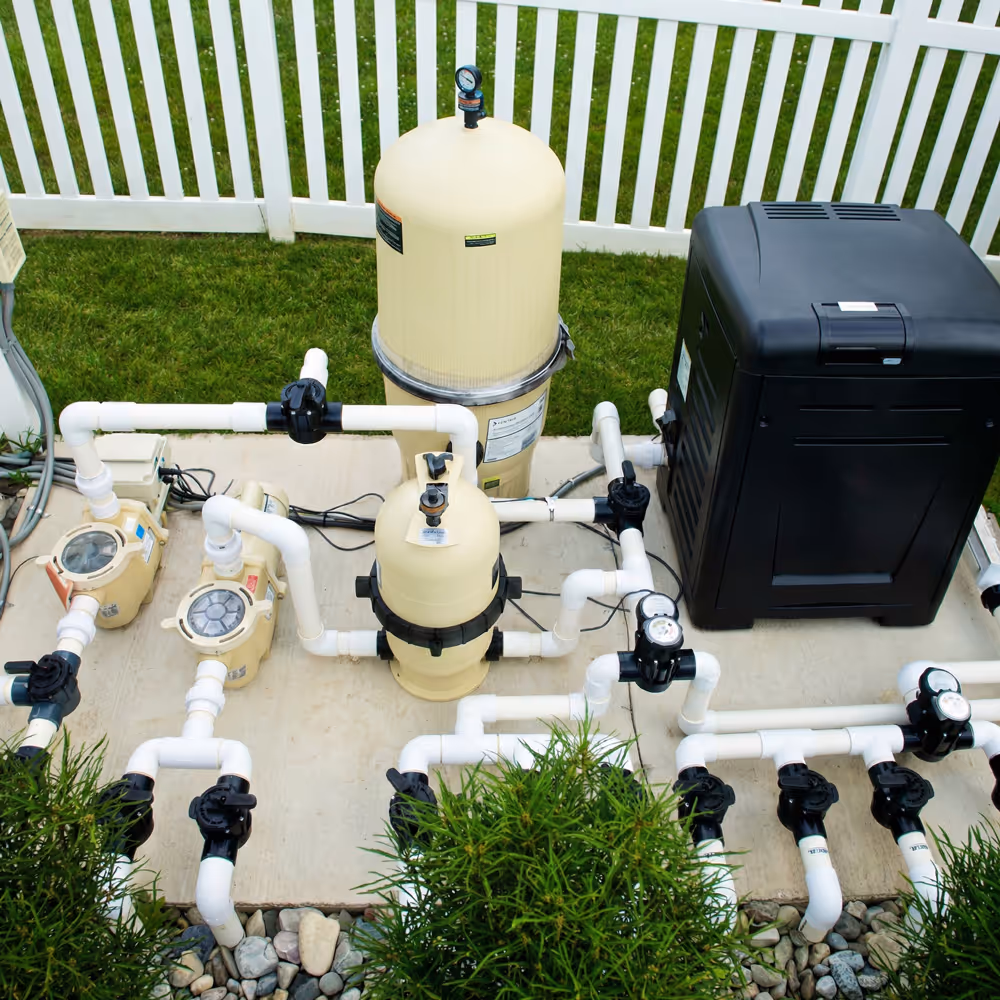 Outdoor pool filtration system with beige tanks, white PVC pipes, gauges, black valves, and a white picket fence in background.