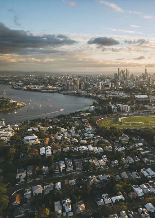 Aerial view of a cityscape with a river, boats, residential area, and skyscrapers at sunset under a partly cloudy sky.