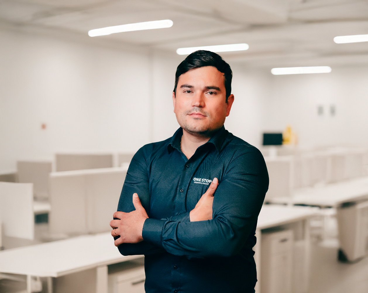 Man with dark hair wearing a navy blue shirt with crossed arms in an indoor setting.