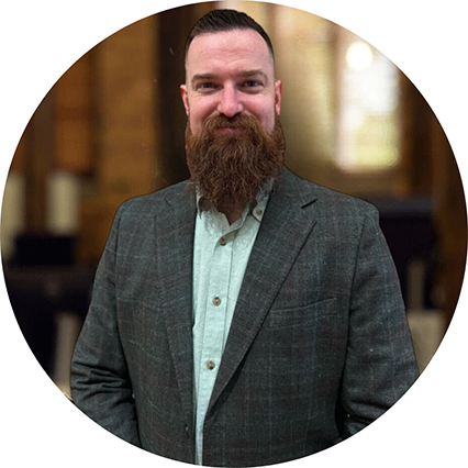 Bearded man wearing a gray checkered blazer over a white shirt standing indoors with a blurred background.
