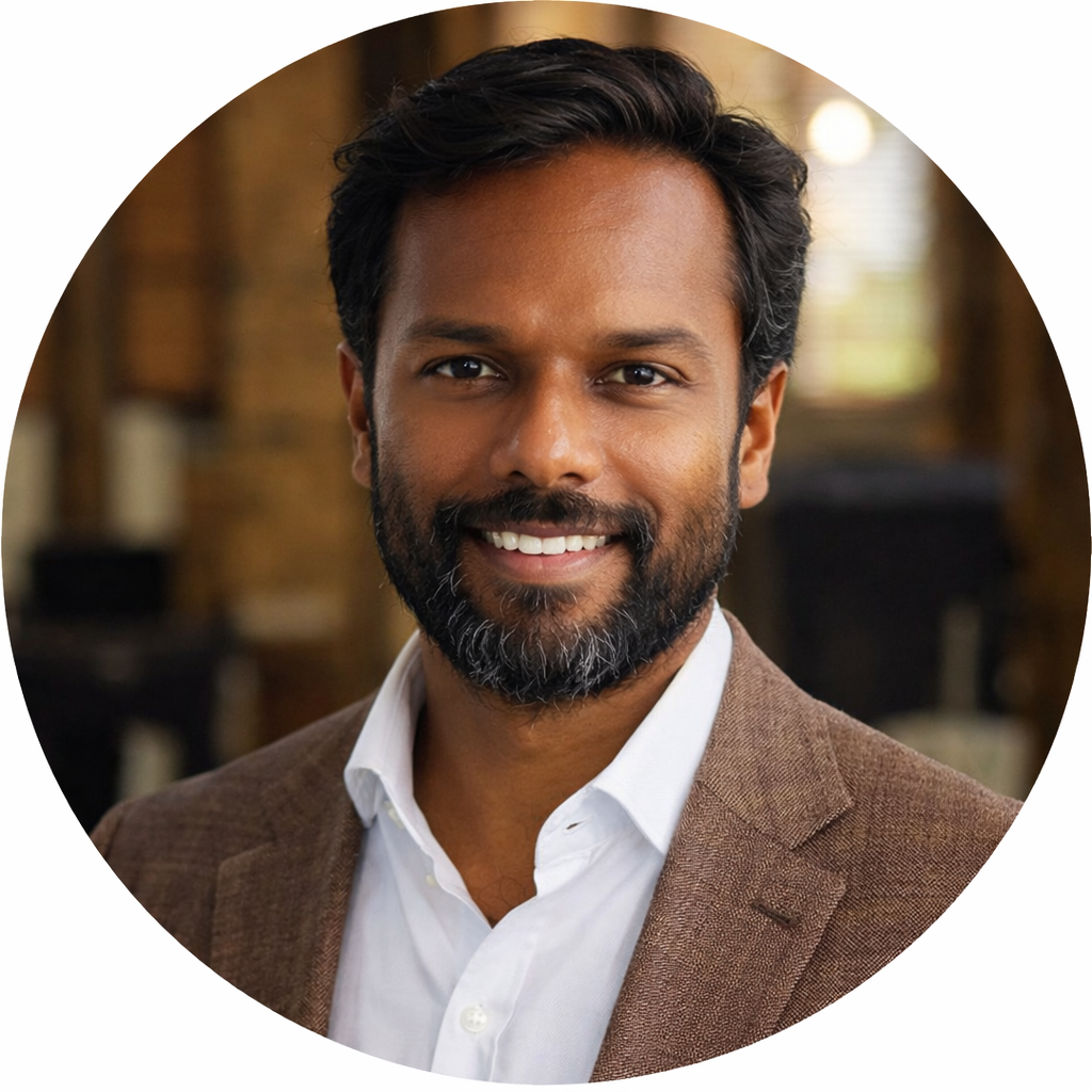 Smiling man with dark hair and beard wearing a brown blazer and white shirt, indoors with blurred background.