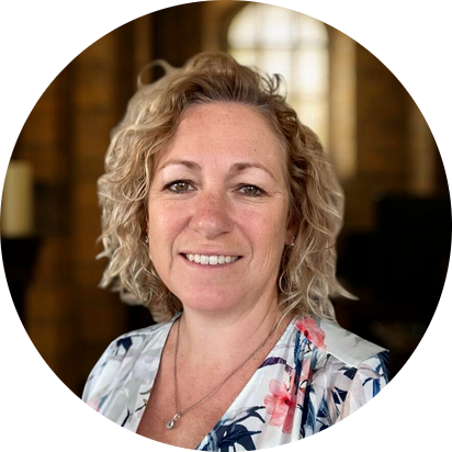Smiling woman with curly blonde hair wearing a floral blouse and a necklace in a softly lit indoor setting.