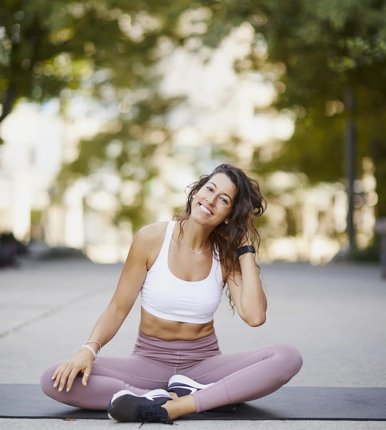 Smiling young woman in white sports bra and mauve leggings sitting cross-legged on a mat outdoors.