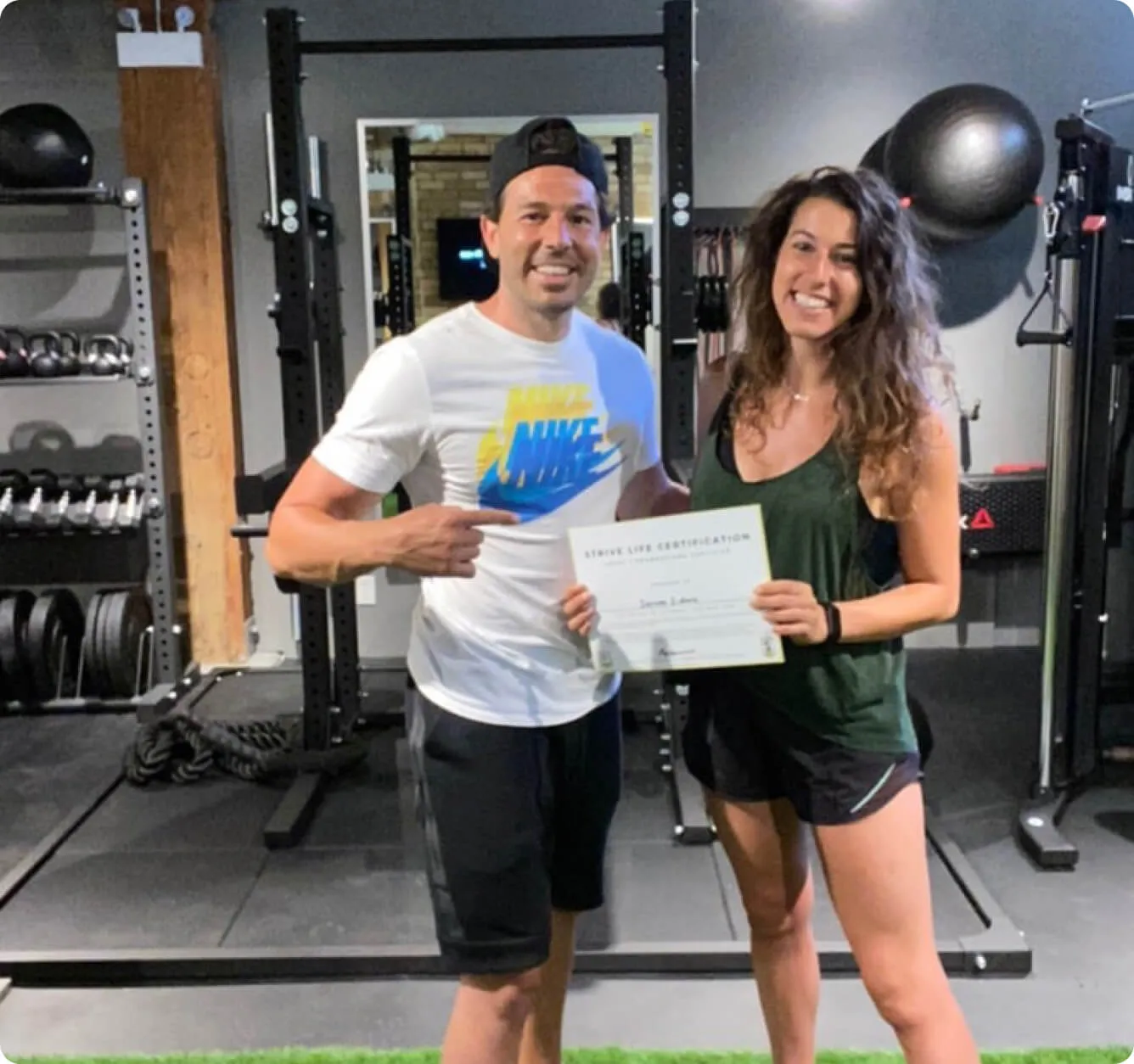 Smiling man in a white Nike shirt and black cap pointing to a woman holding a certificate in a gym.