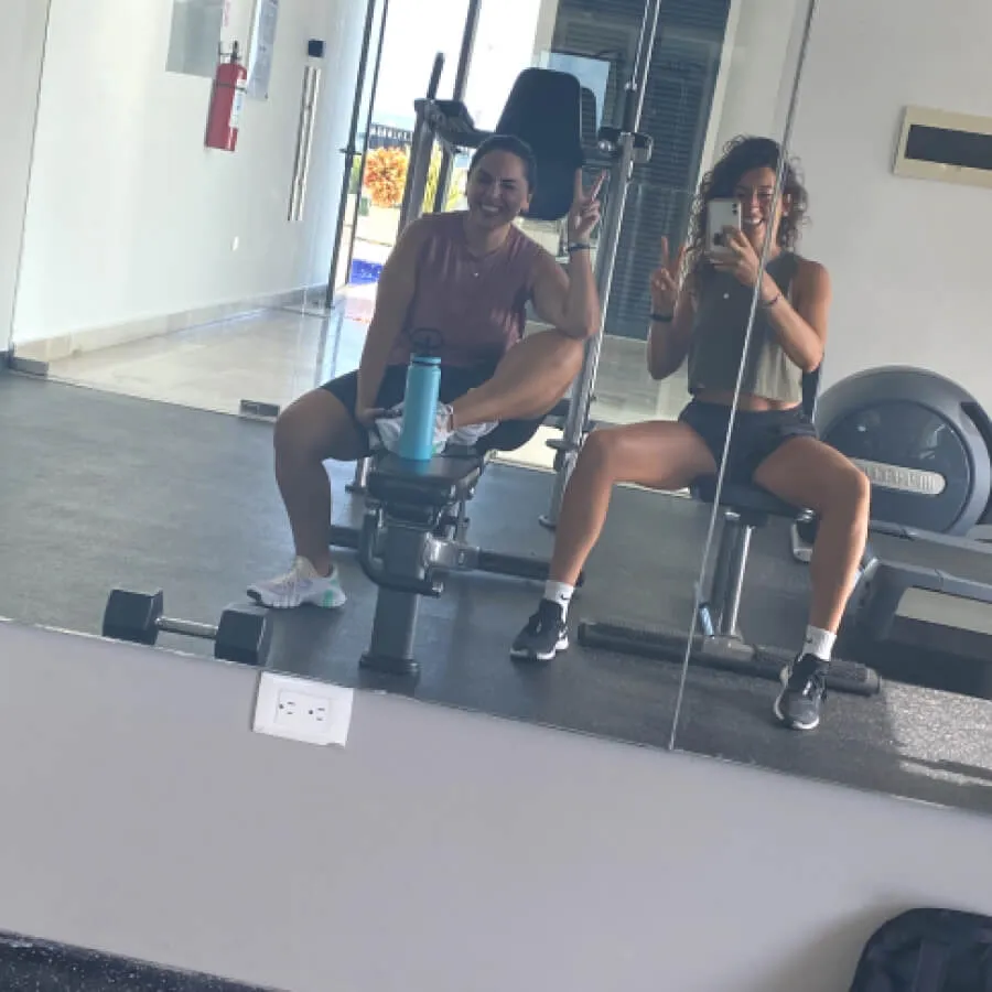 Two women sitting on gym benches, smiling and making peace signs in front of a gym mirror.
